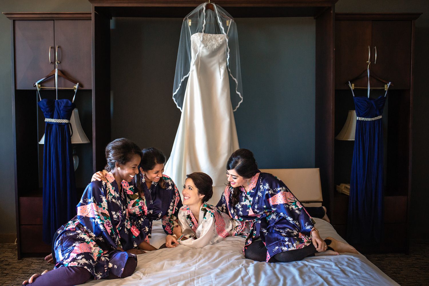 Bridesmaids in floral robes gather on a bed admiring the bride beside a hanging wedding gown.