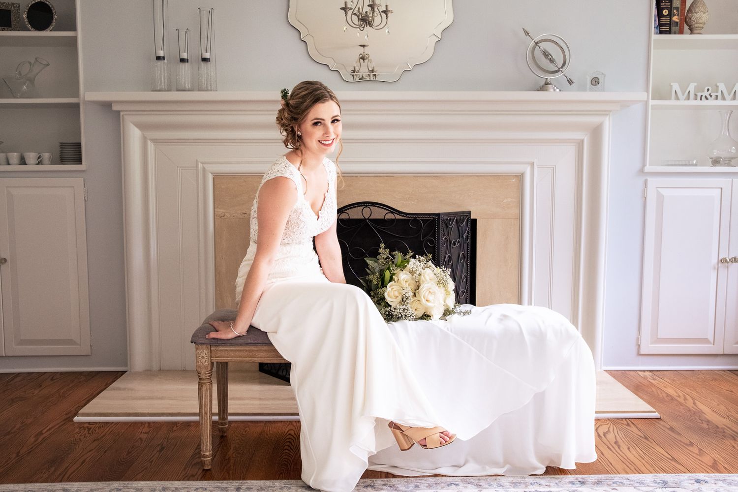 Elegant bride in white gown sits gracefully by a fireplace holding a bouquet.