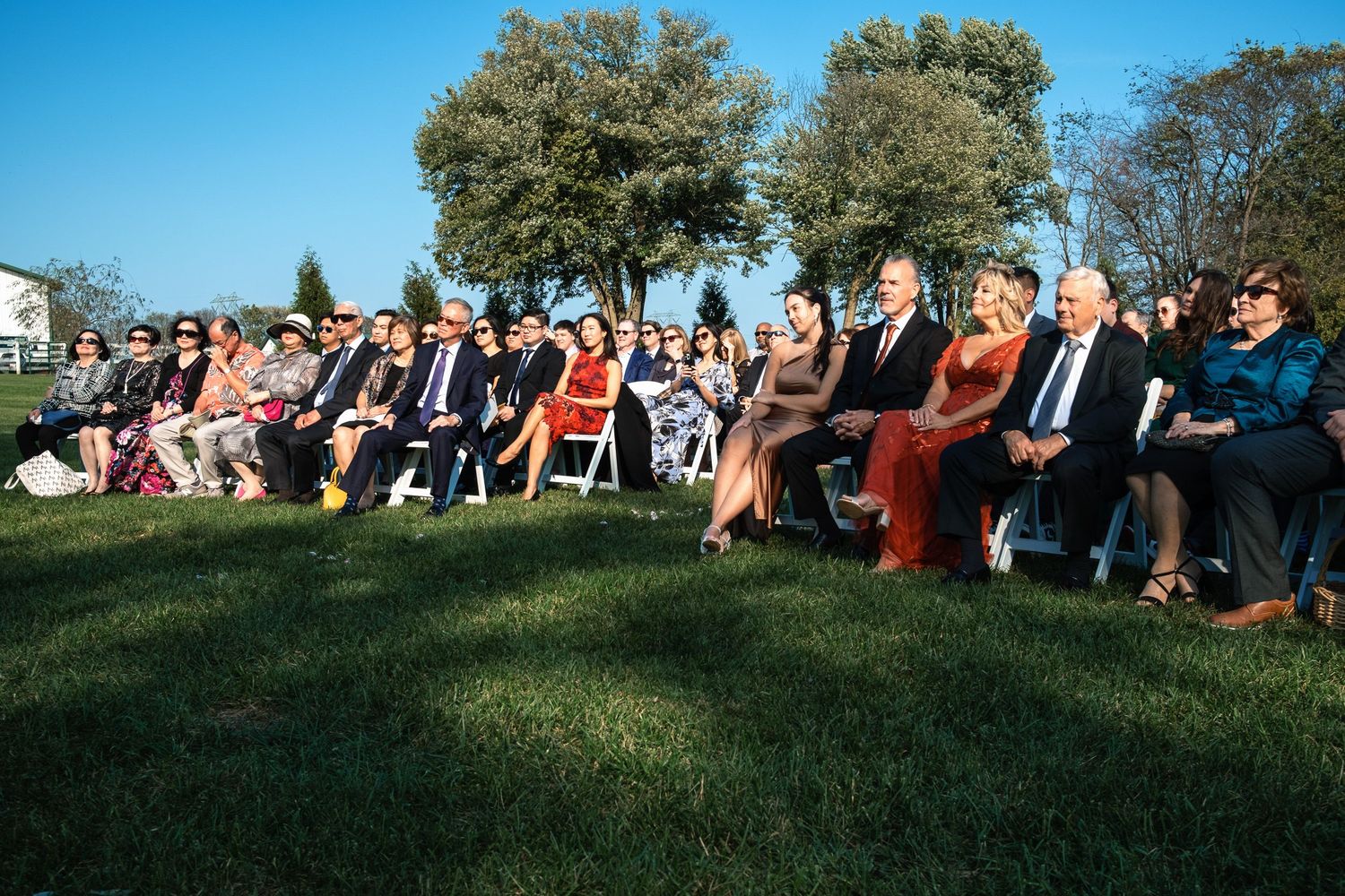 Large group of wedding guests seated on white chairs at an outdoor afternoon farm wedding ceremony.