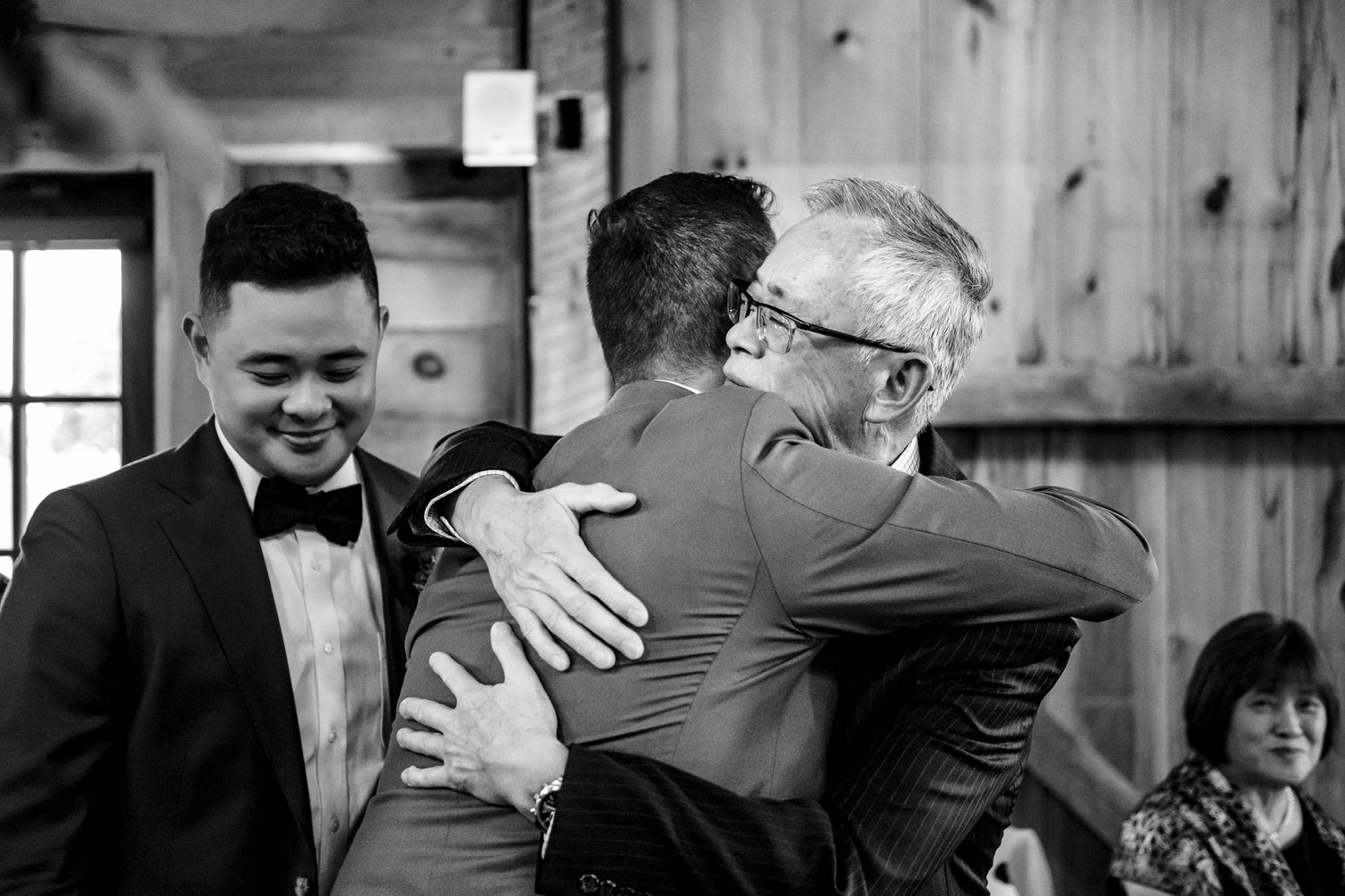 Emotional wedding moment as two men share a heartfelt hug while groom in bow tie watches.