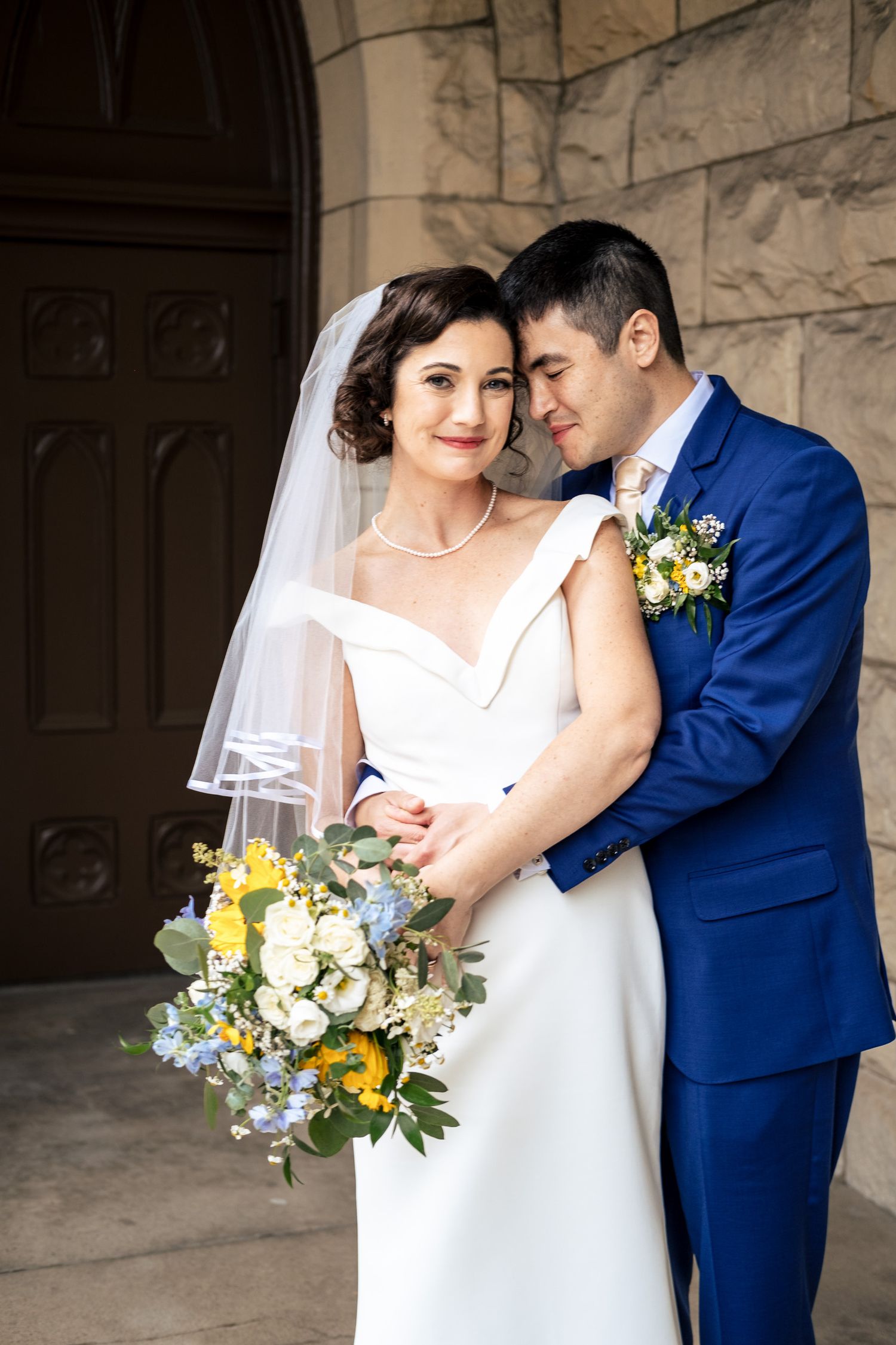 Groom in blue suit embraces bride in white gown holding yellow and blue bouquet outside church.
