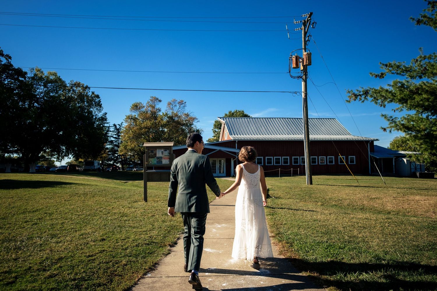 Newlyweds walk hand in hand toward blue barn venue on sunny wedding day.