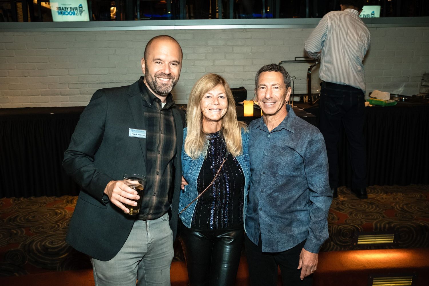 Three smiling adults pose together at an indoor social event, with a bar and guests visible in the background.