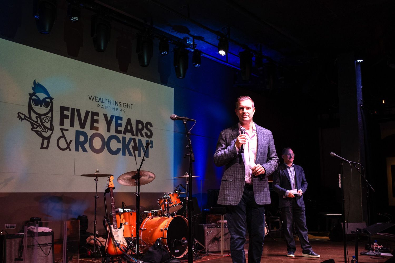 Man speaking on stage at Wealth Insight five year anniversary event with band setup and blue lighting backdrop.
