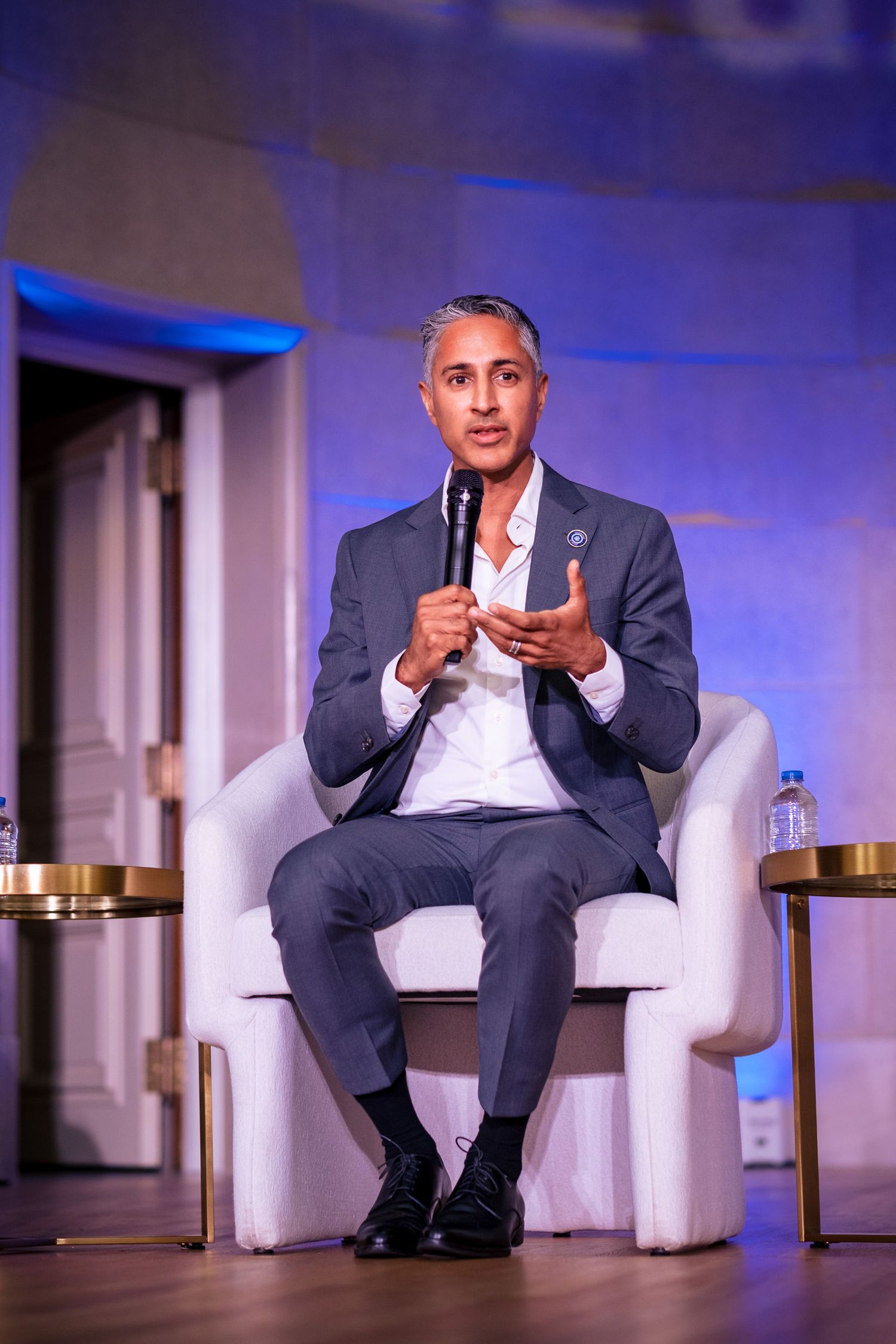 Man in gray suit speaking at panel event, holding microphone while seated in white chair on stage.