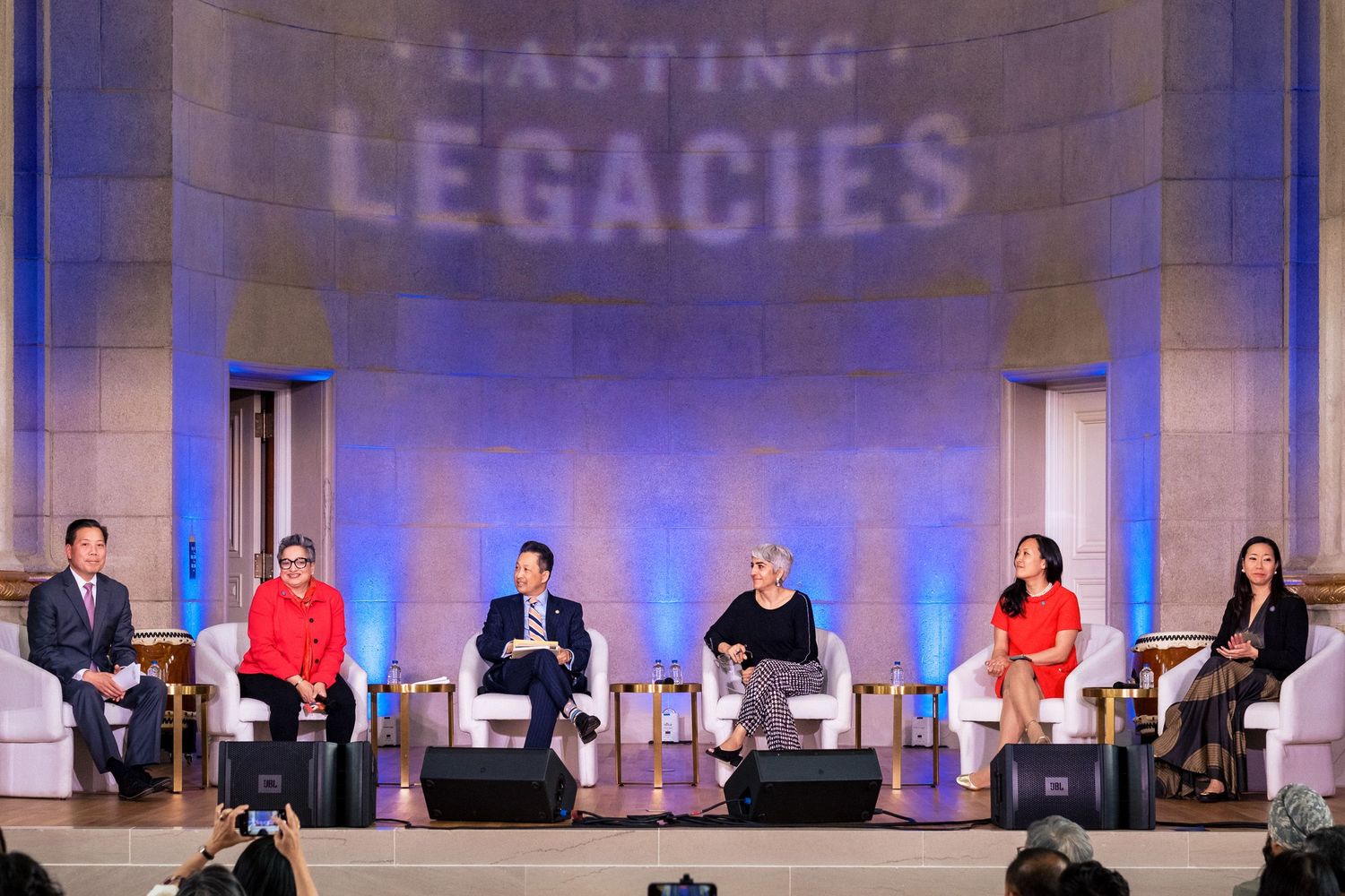 Six panelists seated on stage at a Lasting Legacies event, engaged in a formal discussion under blue lighting.
