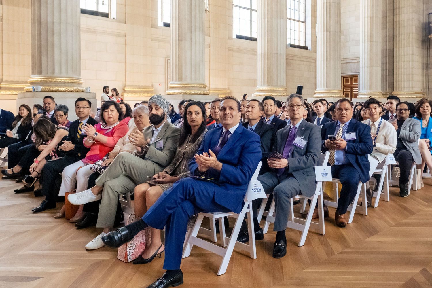 Diverse group of formally dressed attendees seated in rows at an elegant ceremony in a grand hall.