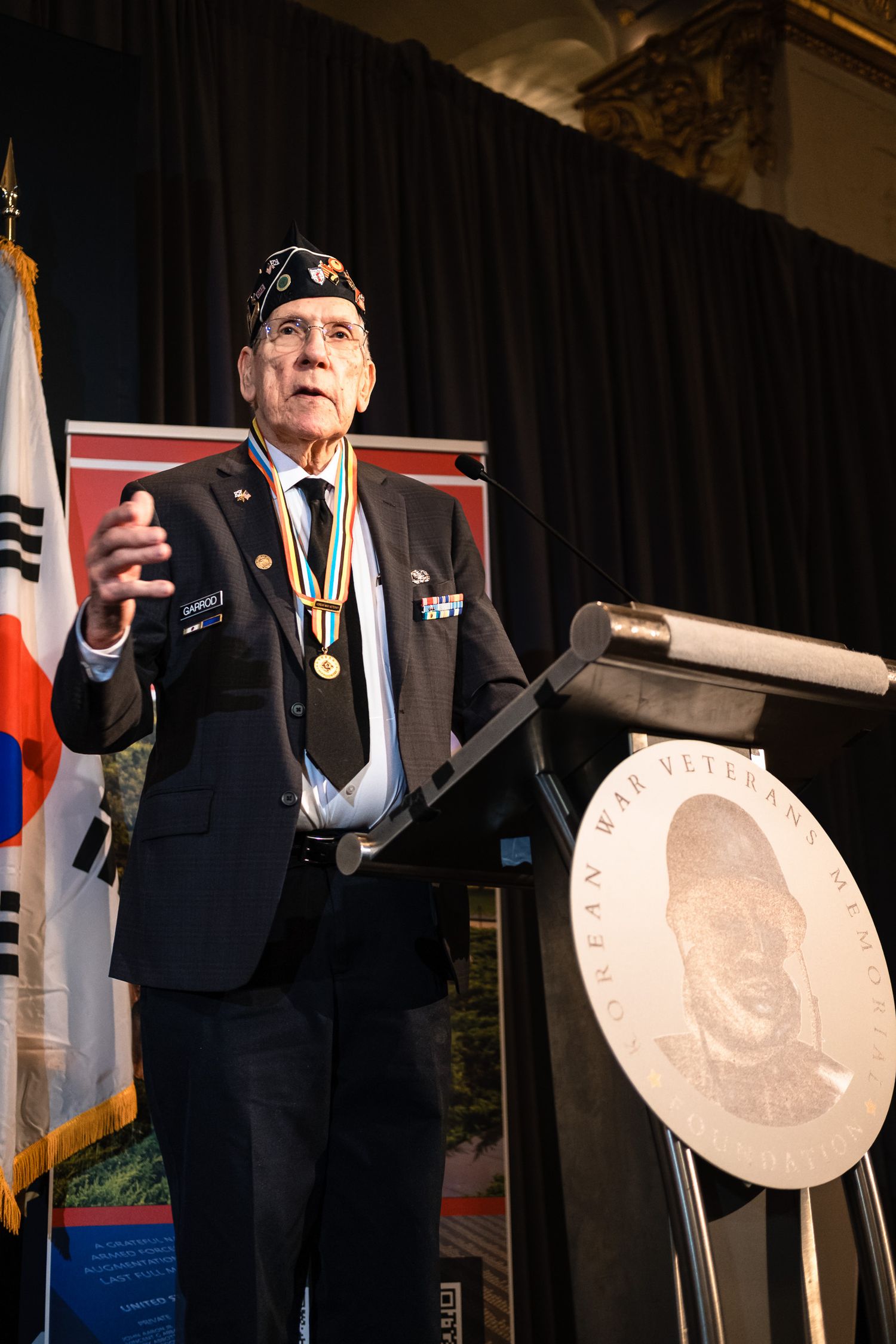 Korean War veteran speaks at podium wearing medals and military cap at commemorative foundation event.