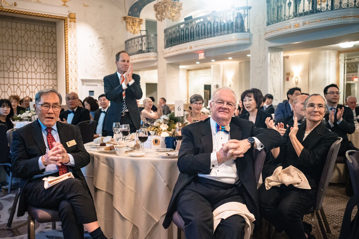 Guests applaud and smile at a formal gala dinner in an elegant ballroom with floral centerpieces.