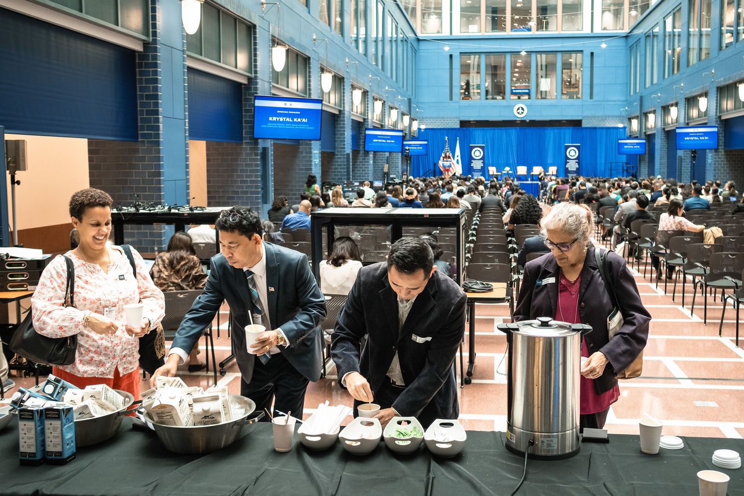 People serving food and beverages at a large indoor event with attendees seated in the background.