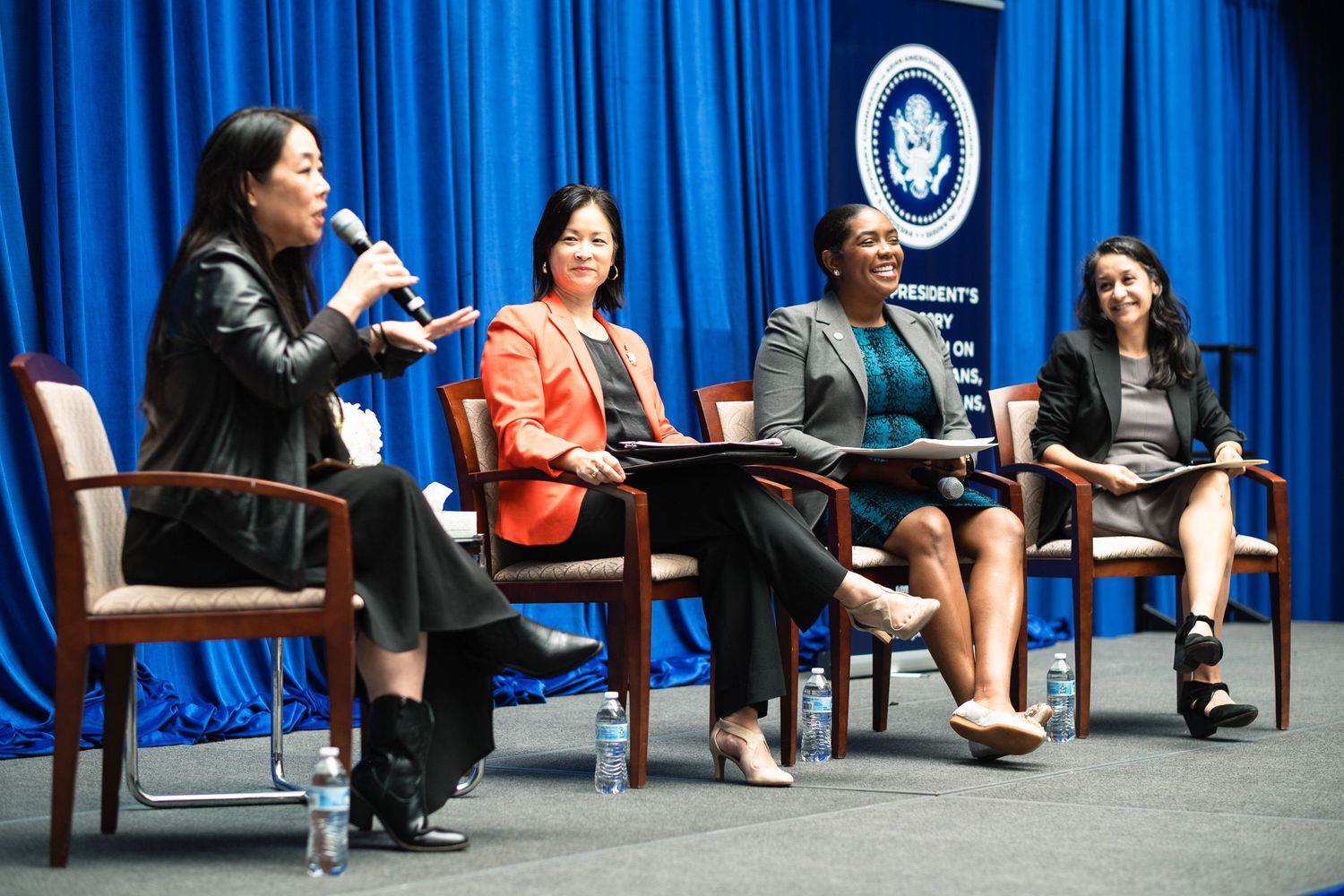 Four women participate in a panel discussion on stage with blue curtains and a presidential seal backdrop.