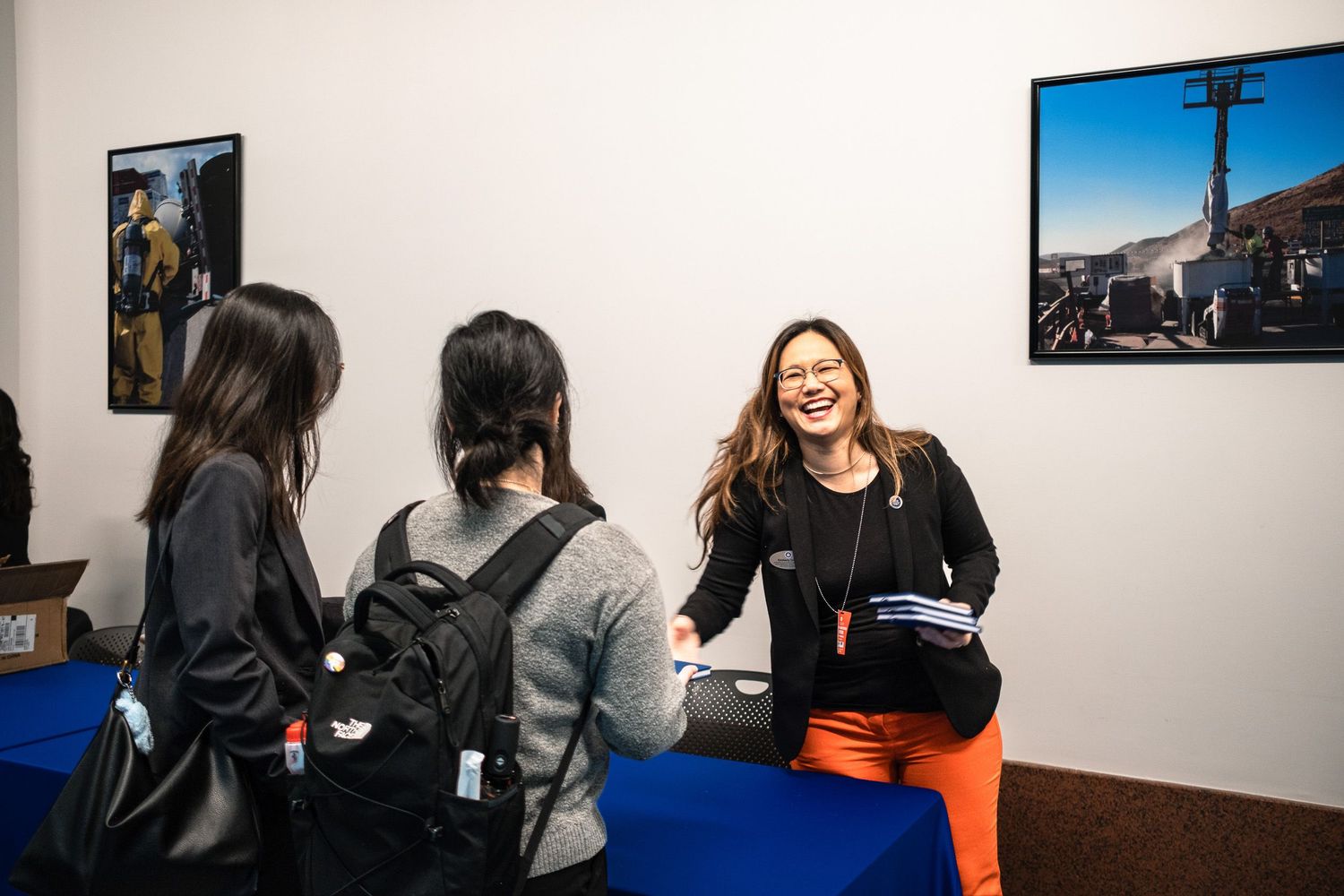 Woman in orange pants smiles at two visitors at a blue table in an art gallery with photos on the wall.