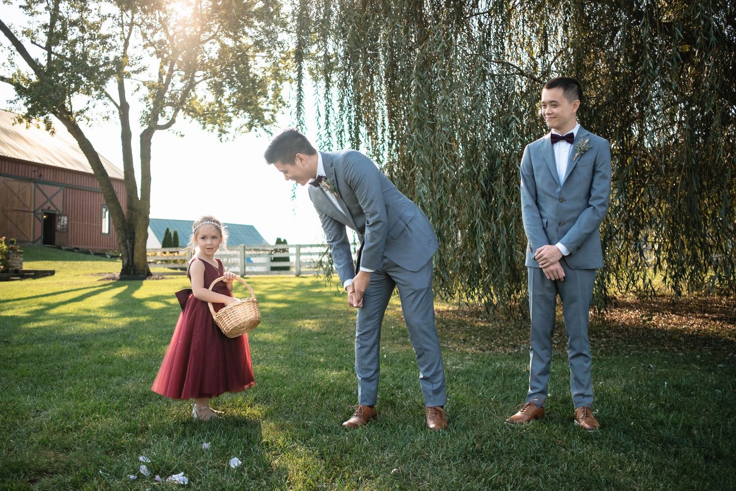 Groomsman in gray suit interacts with flower girl in burgundy dress at outdoor wedding ceremony.