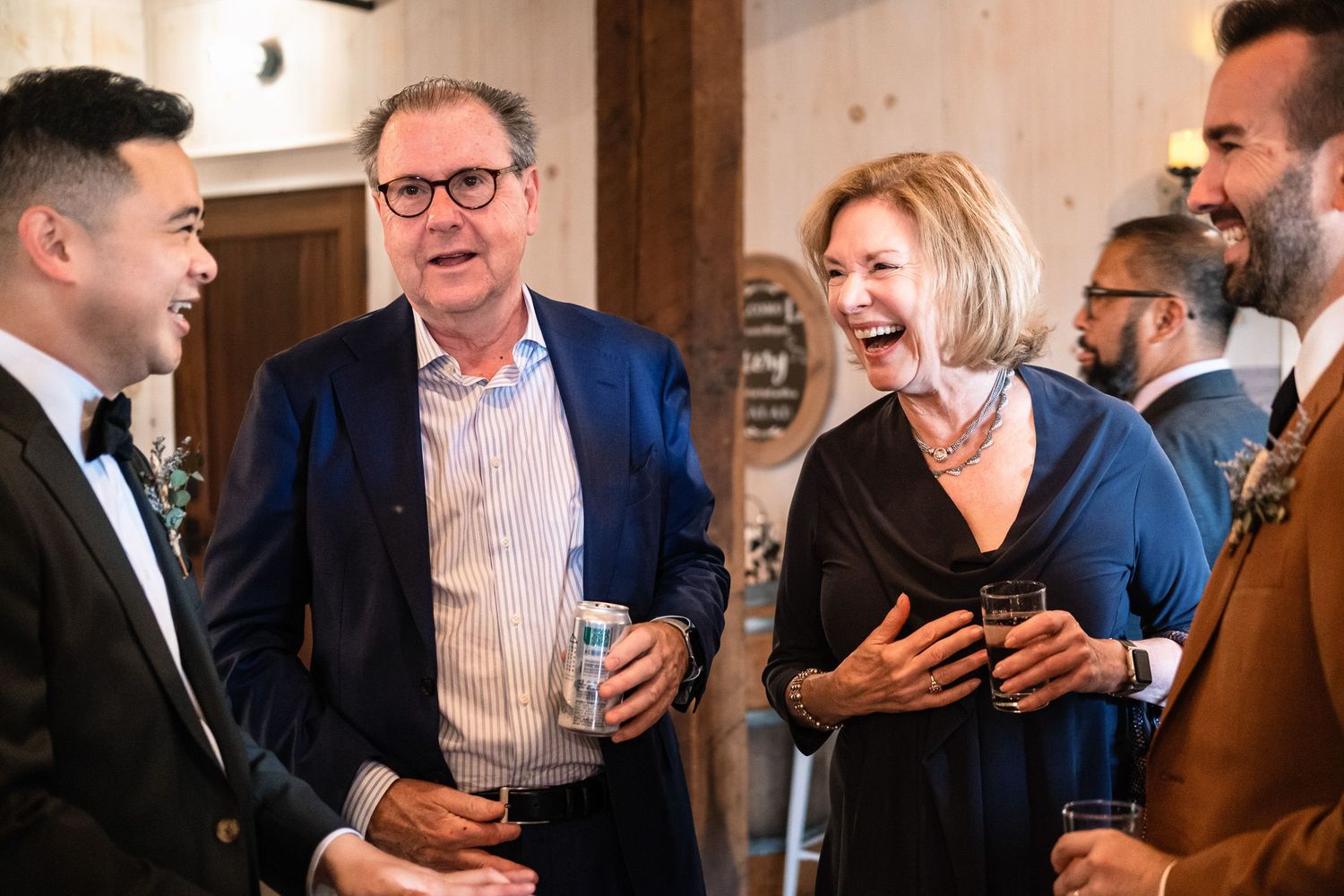 Wedding guests laughing and socializing at rustic barn reception with groom in tuxedo.