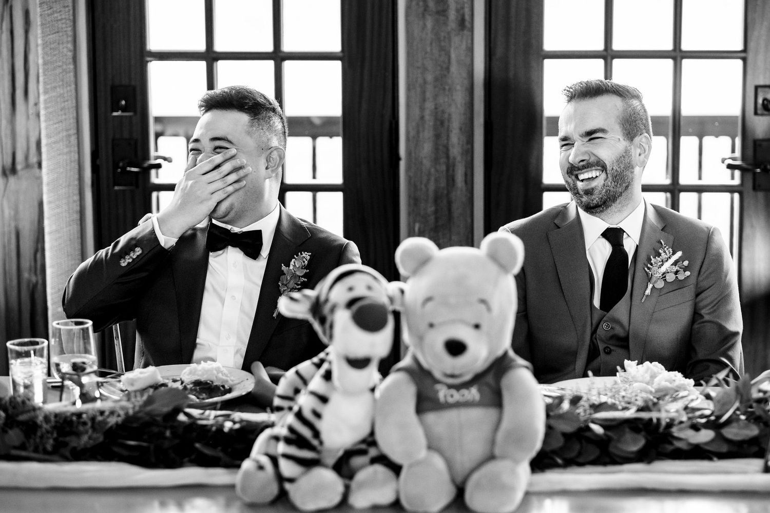Two grooms laugh at reception table with Winnie the Pooh and Tigger plush toys.