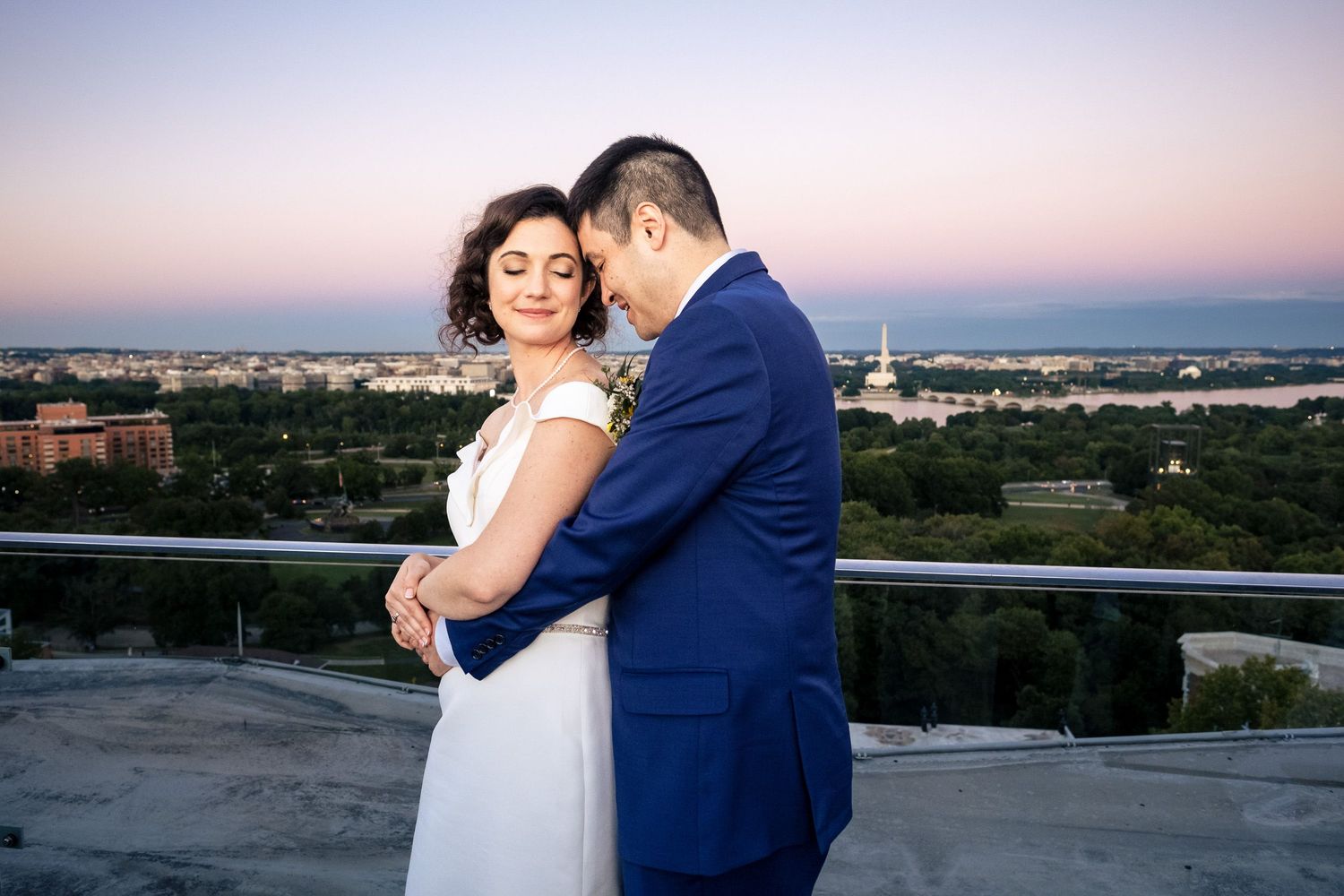 Bride and groom share a romantic embrace on a rooftop at sunset with a city skyline backdrop.