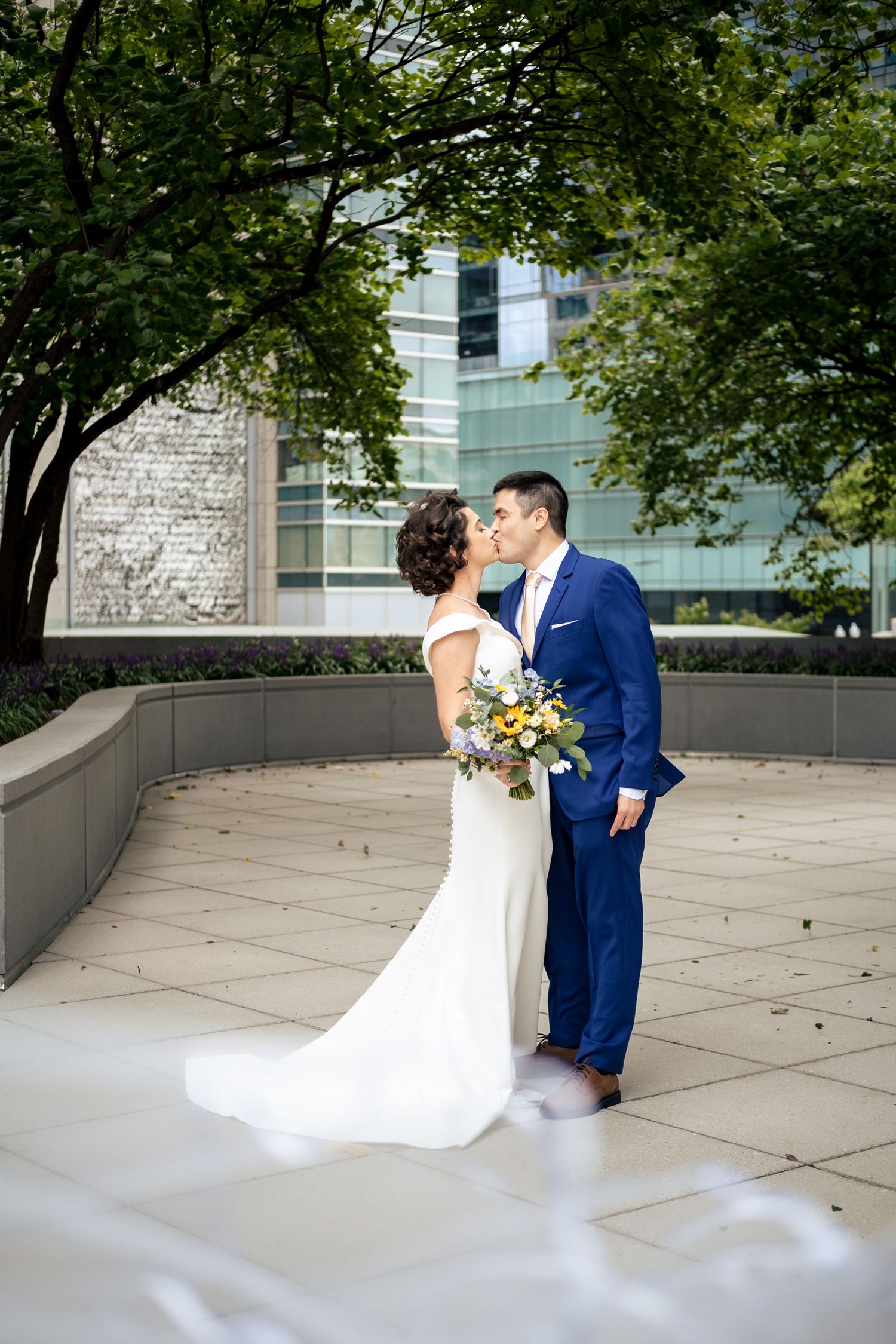 Bride and groom kiss romantically in urban courtyard surrounded by lush green trees on wedding day.
