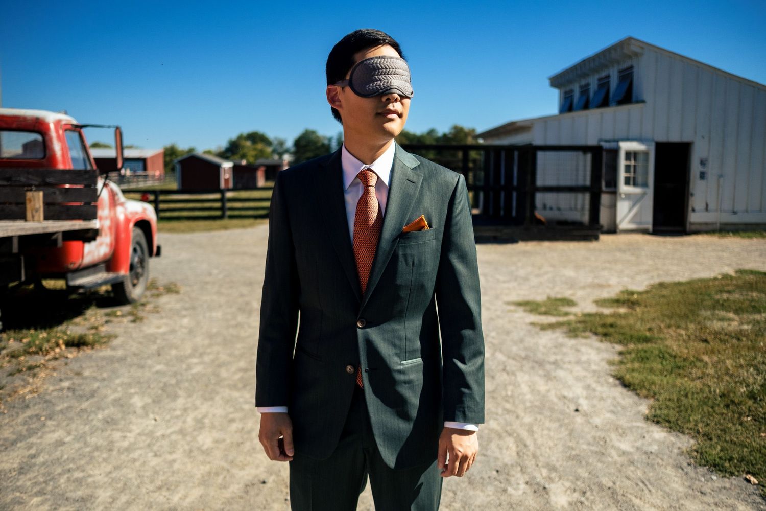 Blindfolded groom in dark green suit standing outside red barn on sunny wedding day.