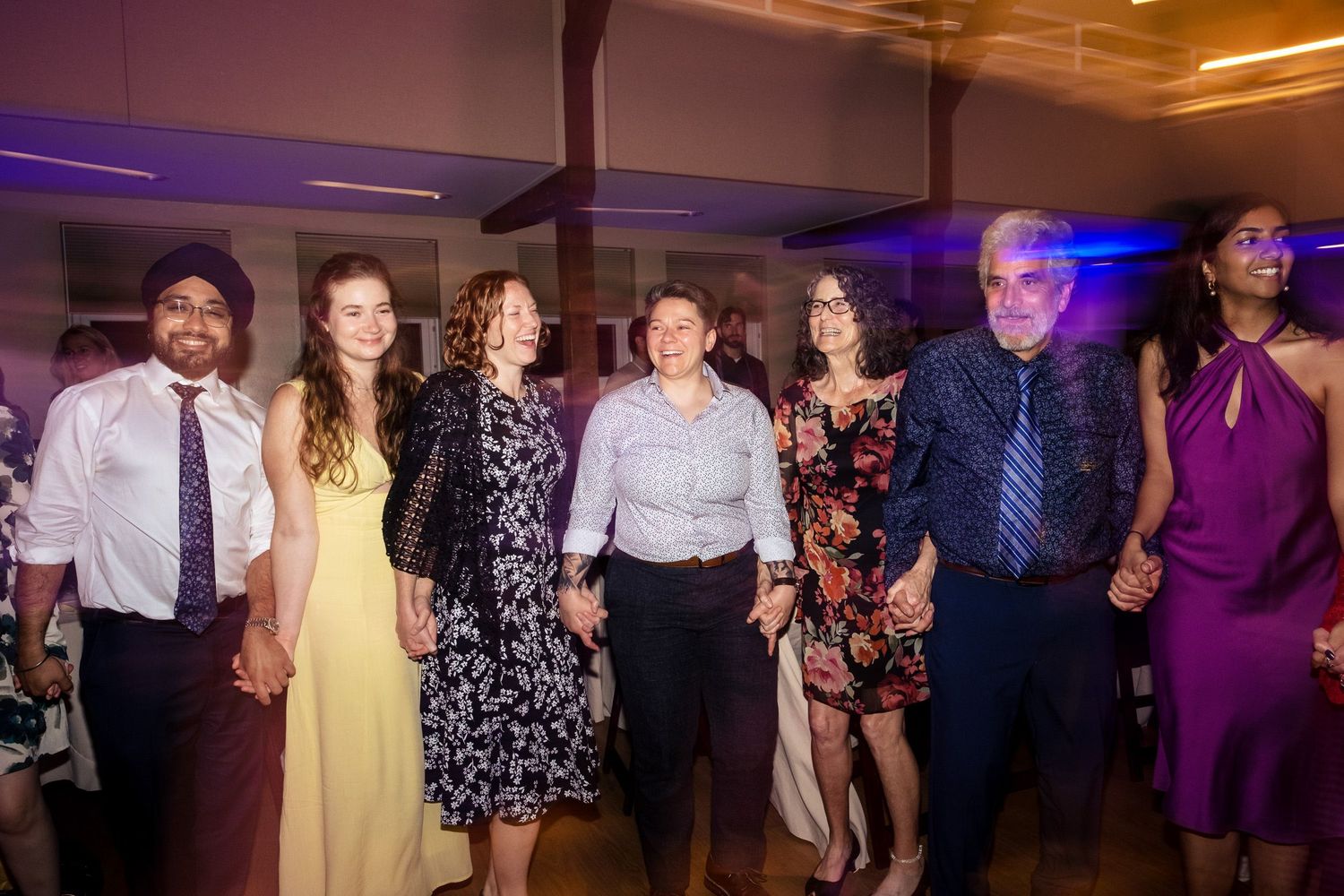 Group of seven wedding guests smile and hold hands together on the reception dance floor.