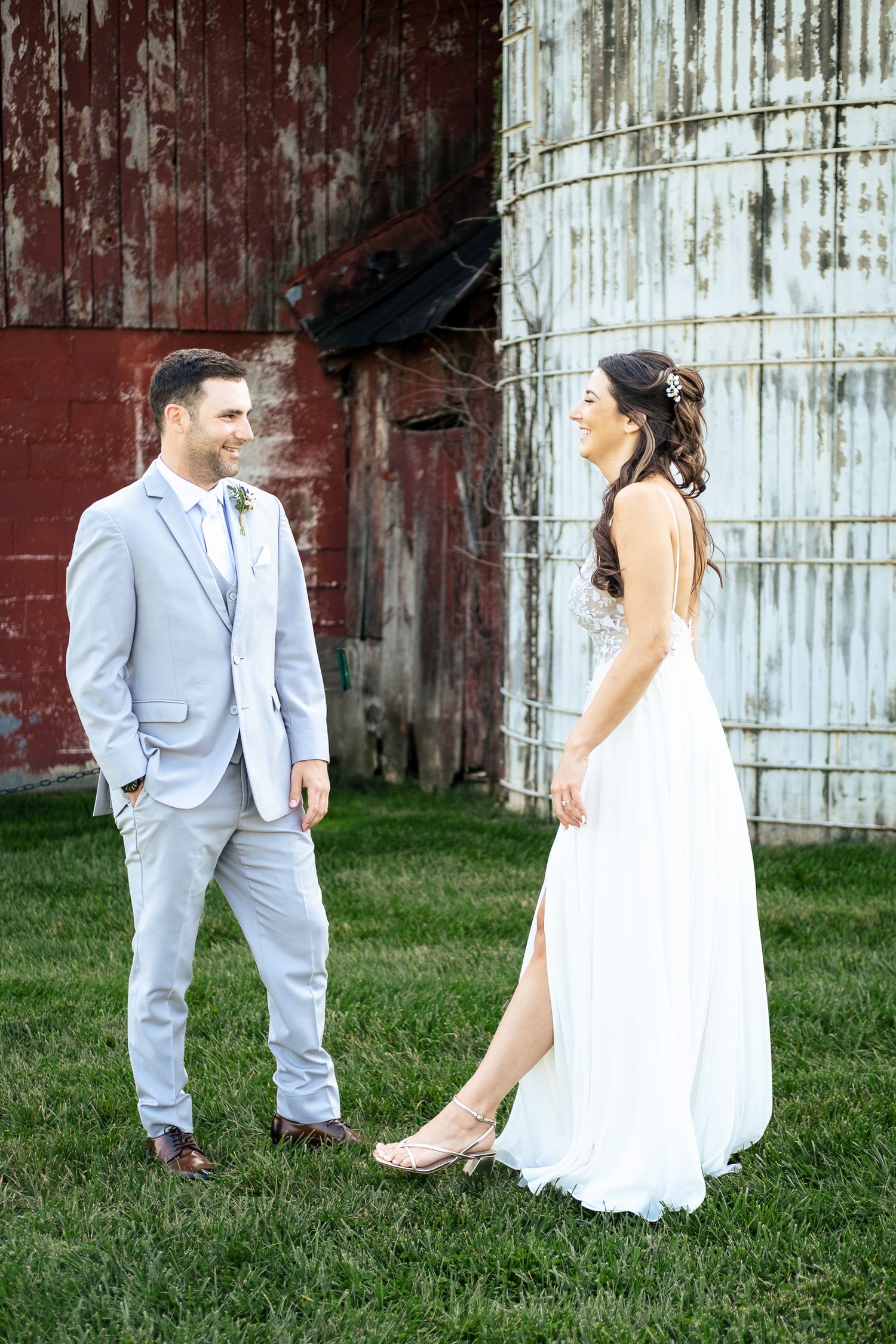 Smiling groom in gray suit and bride in white gown stand together beside rustic red barn.