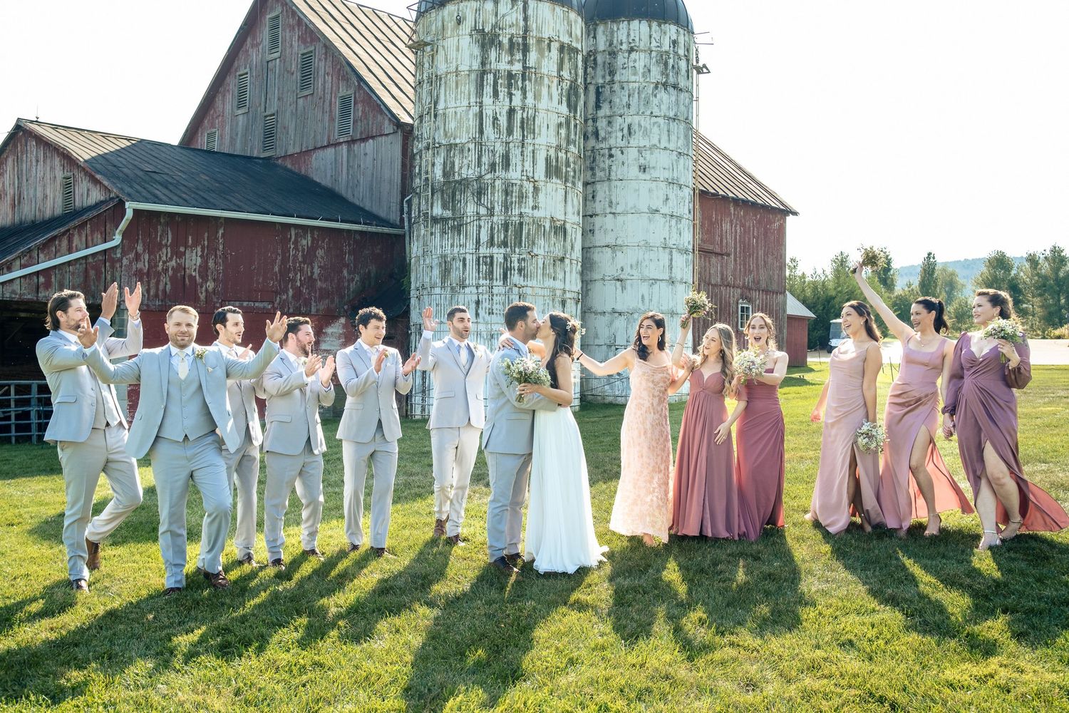 Full wedding party celebrates joyfully on green lawn beside a red barn and grain silos.