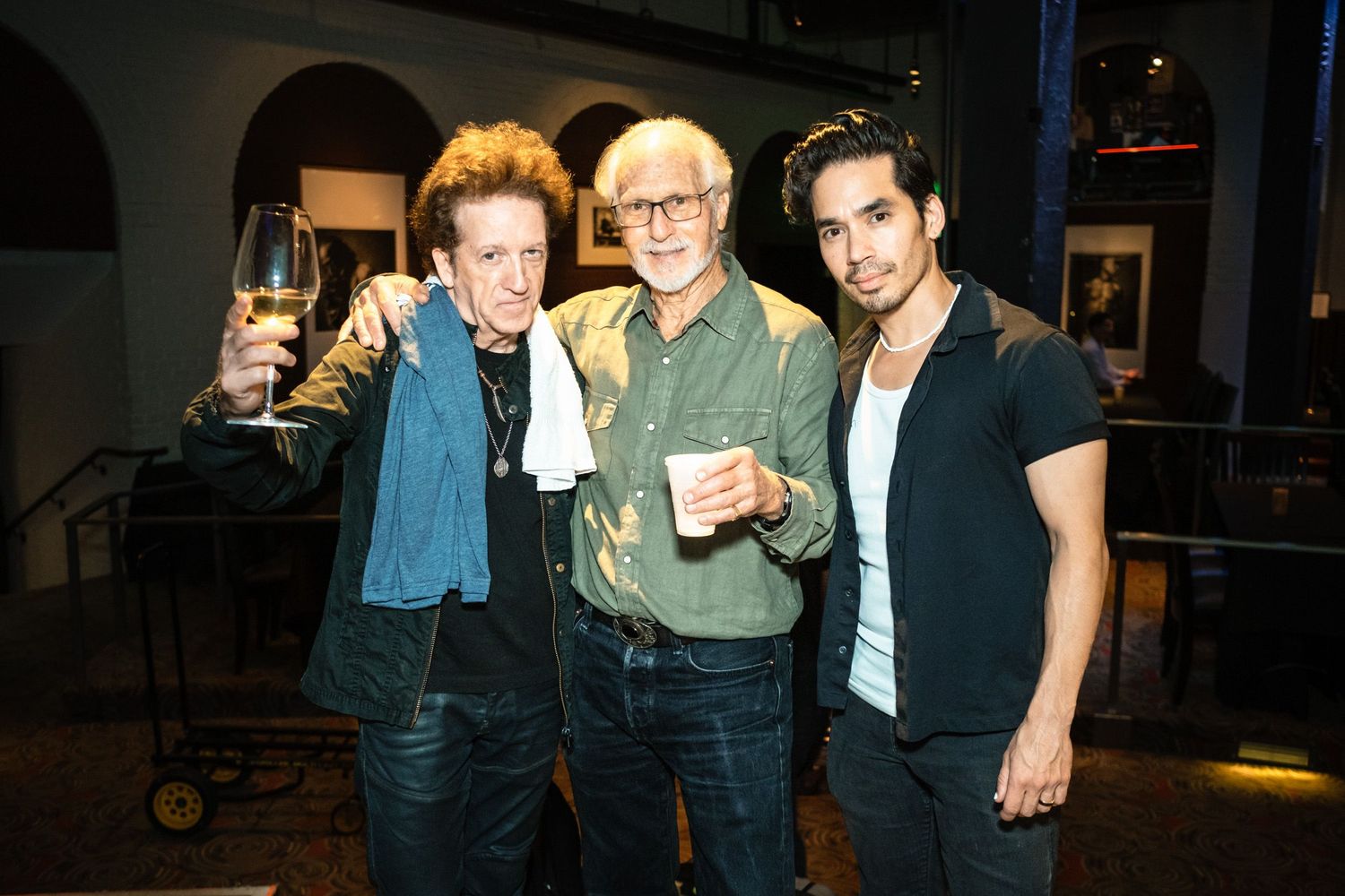 Three men smiling and raising drinks at an indoor event, posing together for a photo in a dimly lit venue.