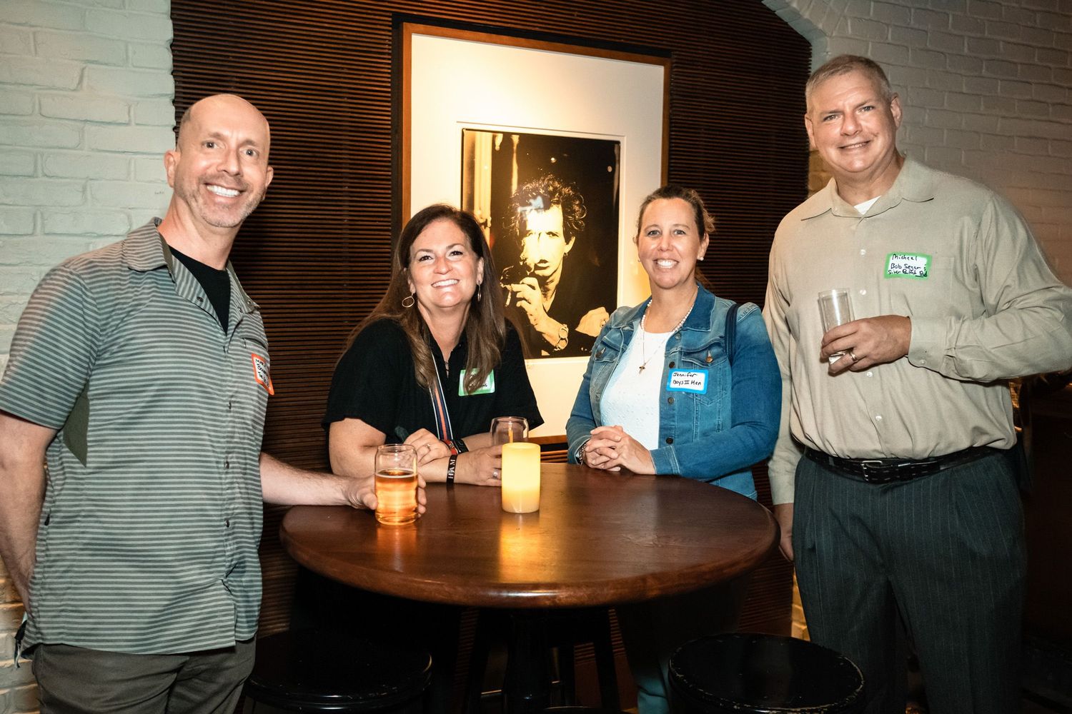 Four adults pose together smiling at a round table with drinks at an indoor social event.