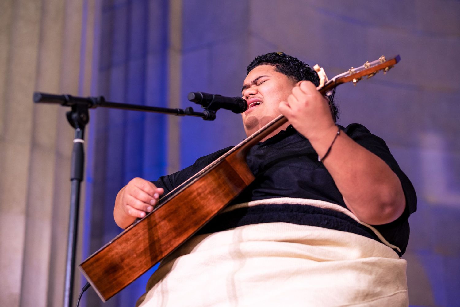 Young musician playing acoustic guitar and singing into microphone on stage during live performance.