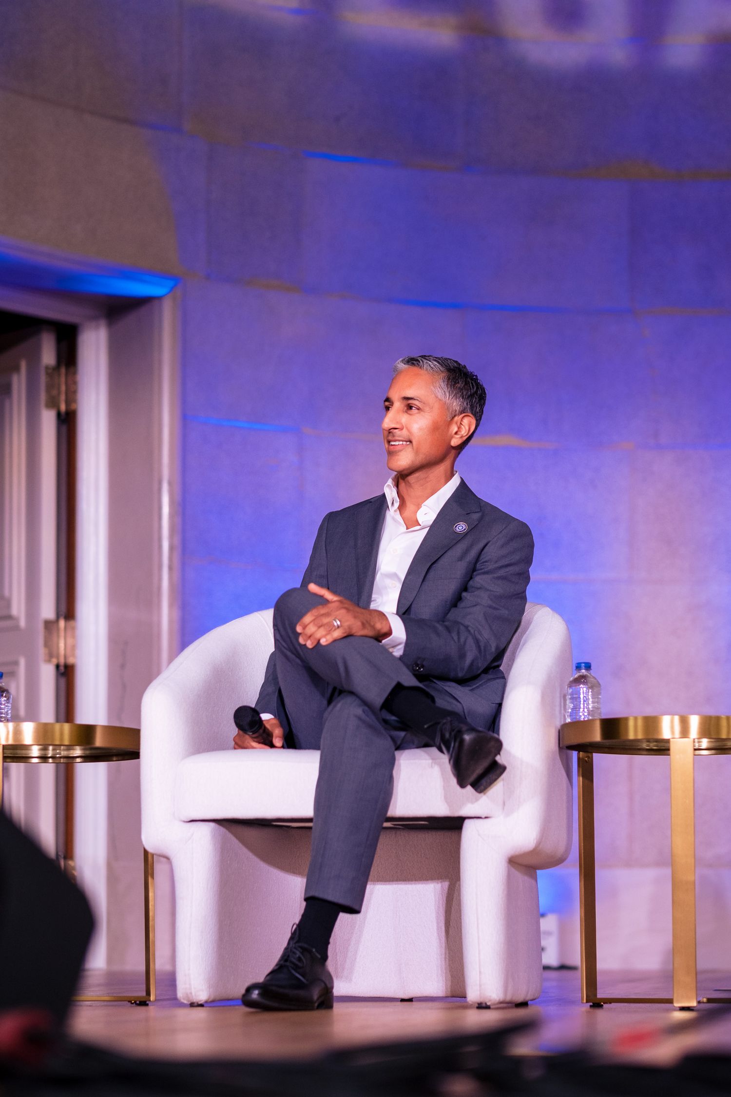 Man in gray suit seated in white chair on stage speaking at professional conference event.