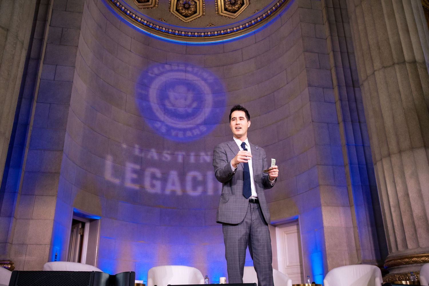 Speaker presents on stage at a Lasting Legacies event inside an elegant venue with blue projected logo.