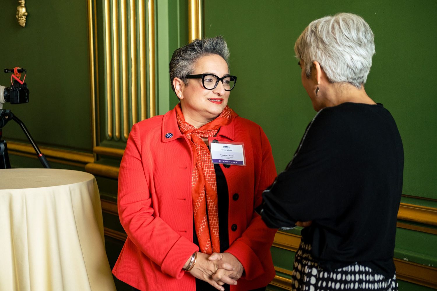 Two women in formal attire engage in conversation at an elegant green-walled event venue.