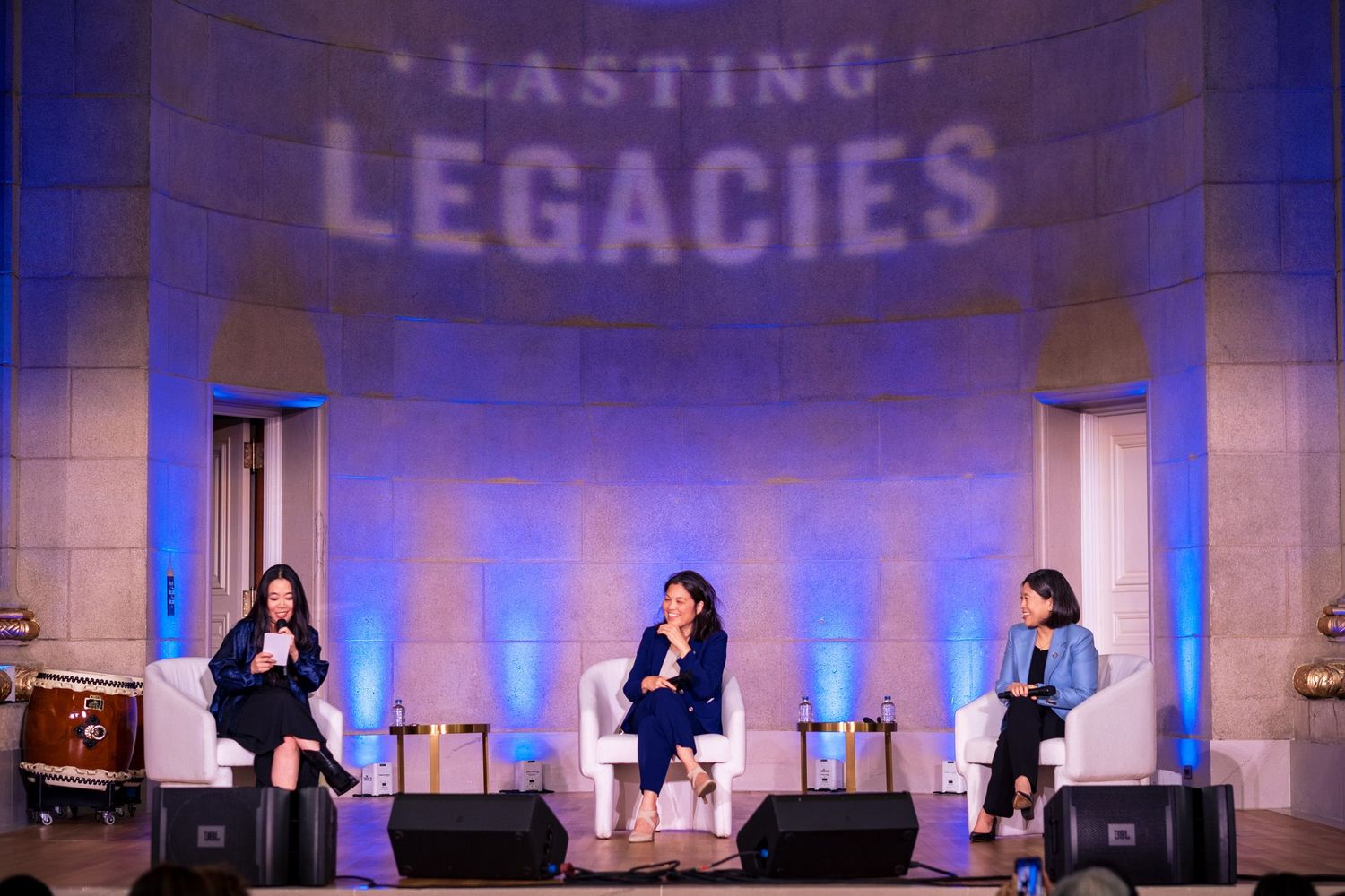Three women on stage at Lasting Legacies event, seated in white chairs under blue lighting.