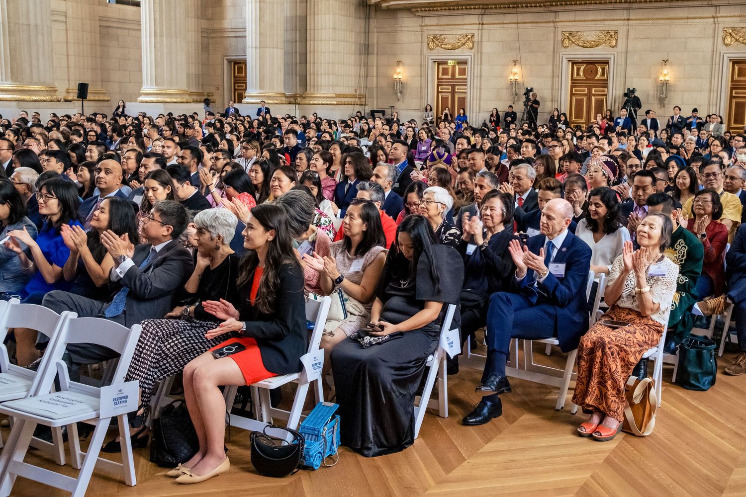 A diverse crowd of attendees applauds at a formal indoor event held in an elegant ballroom.