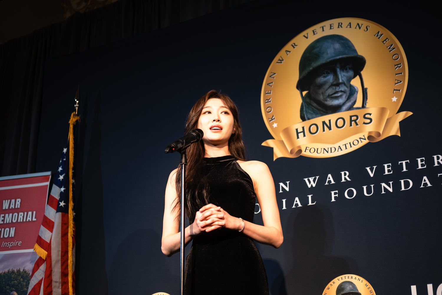 Woman in black dress speaking at Korean War Veterans Memorial Foundation Honors event podium.
