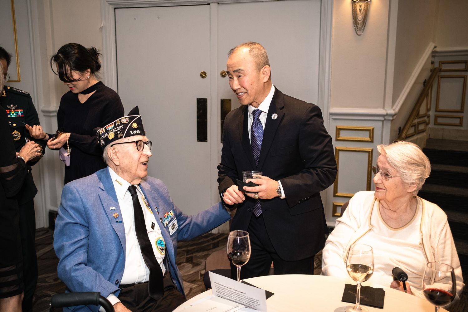 Veteran in blue blazer shaking hands with dignitary at formal event, elderly woman seated nearby with wine glass.