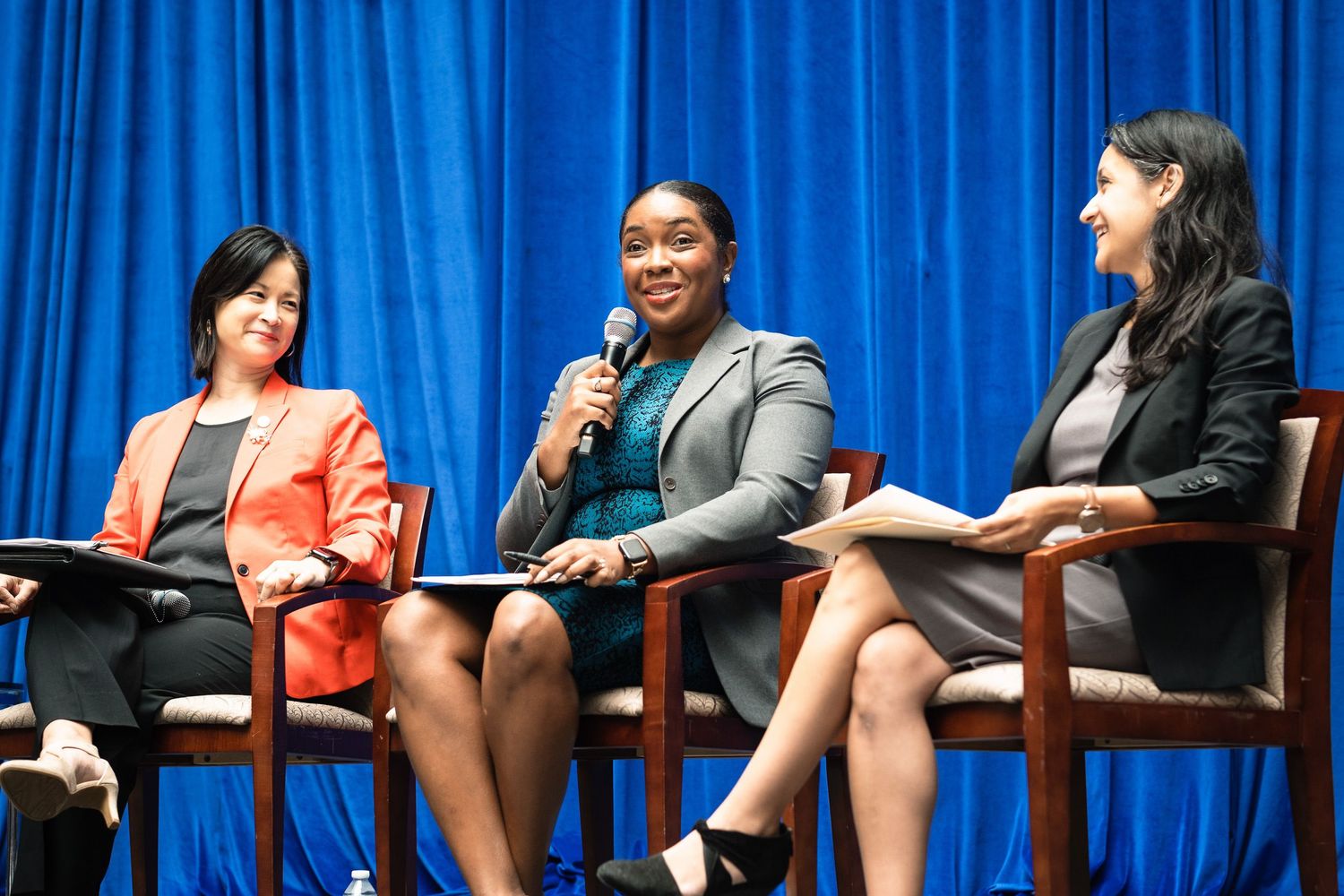 Three professional women seated on stage during a panel discussion in front of a blue curtain backdrop.