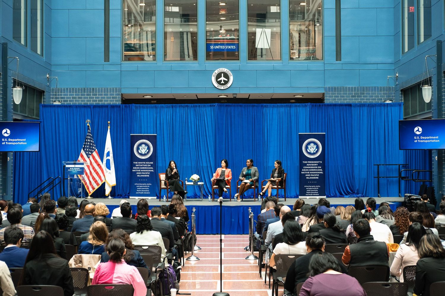 Panel discussion at a government agency event with speakers on stage before an audience, American and organizational flags displayed.