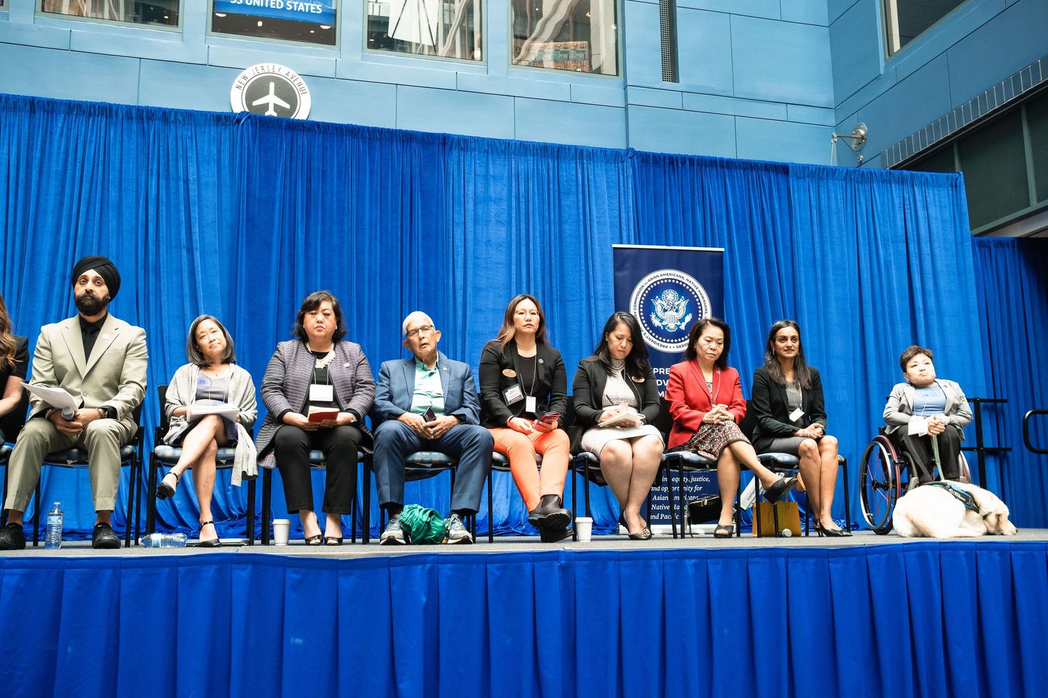 Diverse group of professionals seated on a blue-draped stage at a formal diplomatic or government ceremony event.