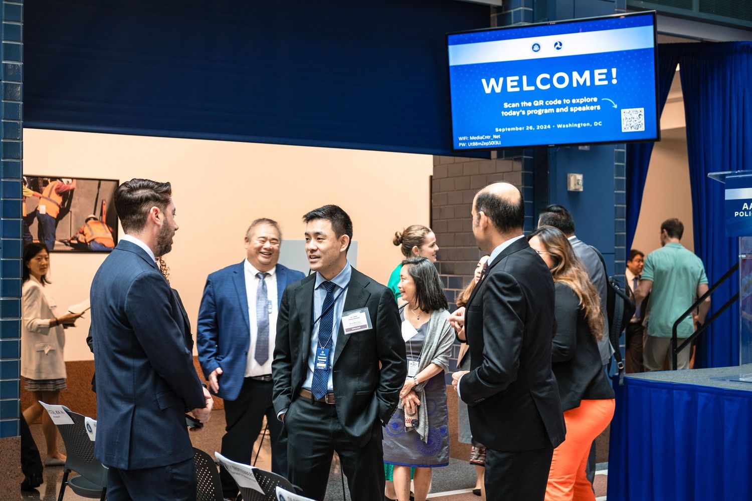 Professionals networking at a welcome reception event with a large WELCOME screen displayed in the background.