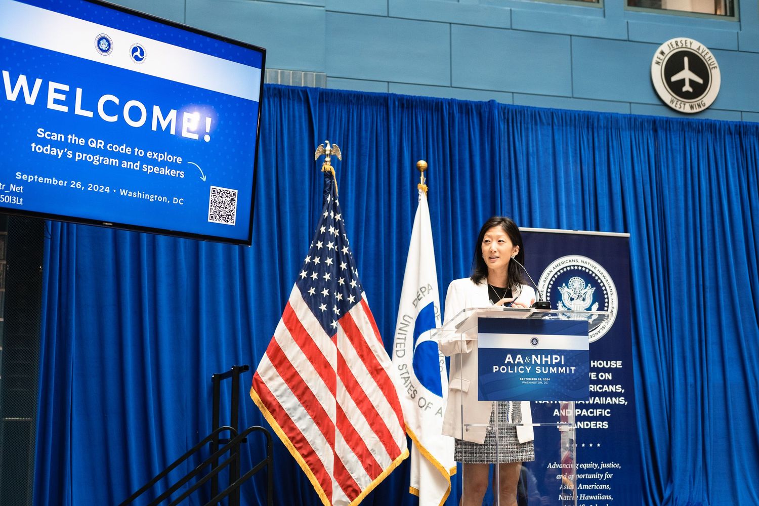 Woman speaking at podium during AANHPI Policy Summit in Washington DC with US flag and welcome screen.