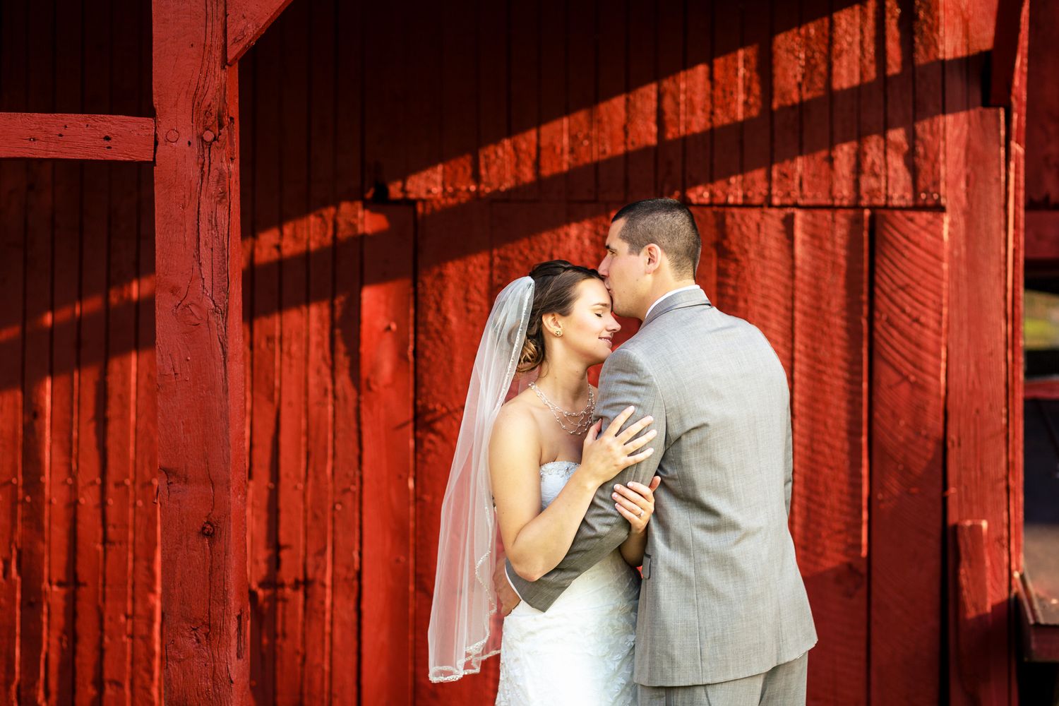Bride and groom share a romantic kiss against a rustic red barn wall in warm sunlight.