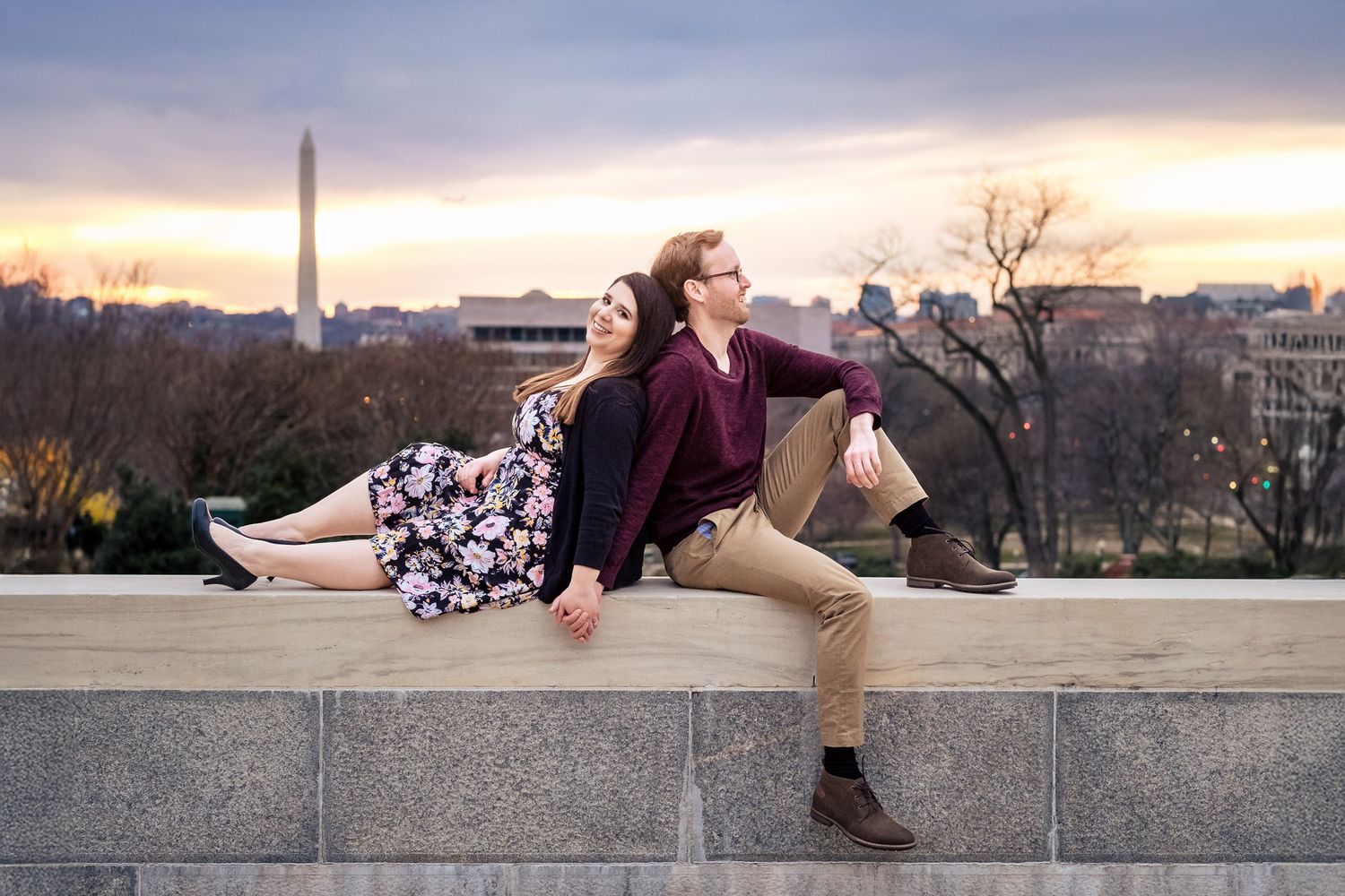 Couple sitting back-to-back on a stone wall with a stunning Washington DC sunset skyline view.