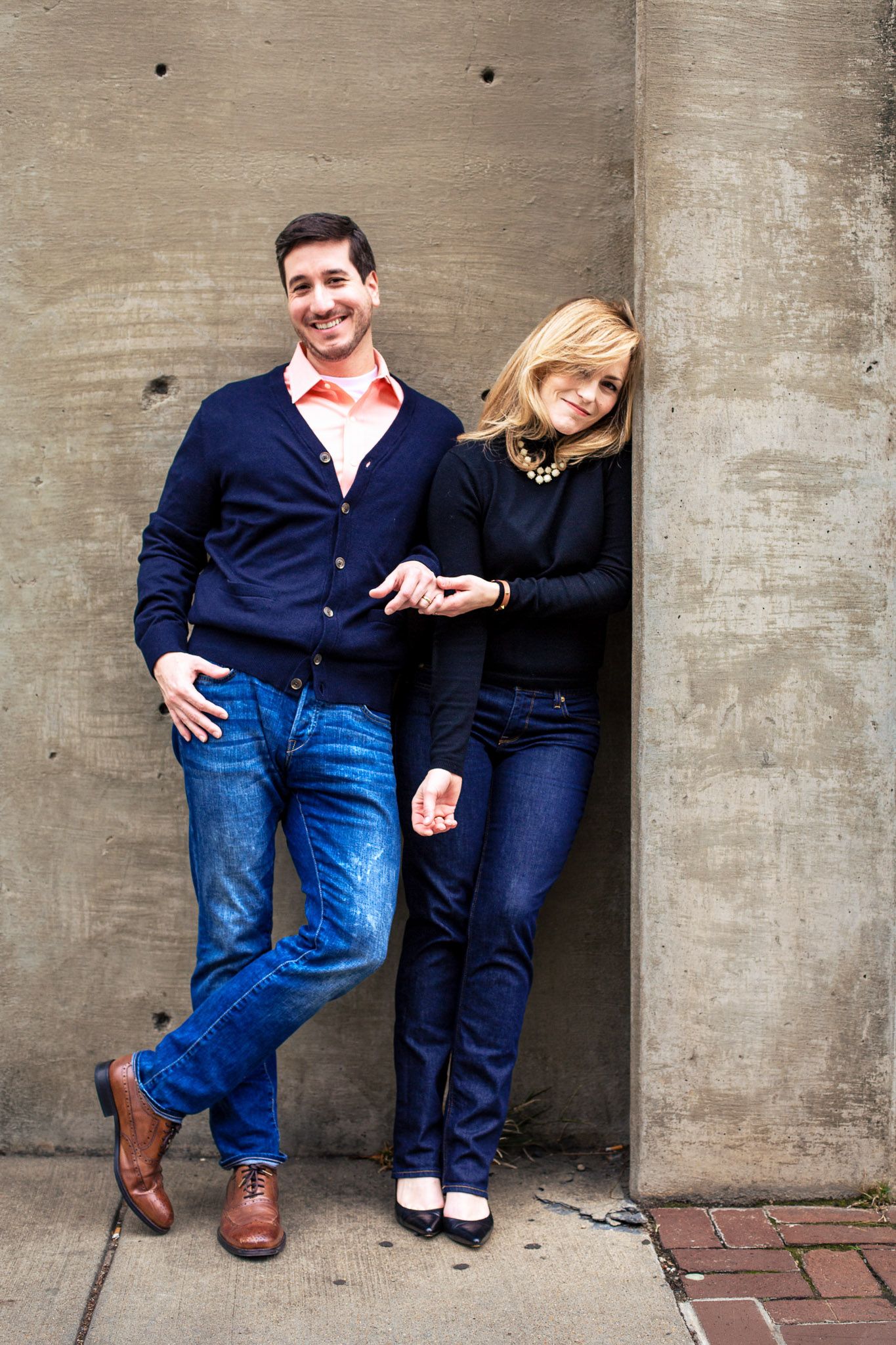 Smiling couple leaning against a concrete wall outdoors, man in navy cardigan and woman in black top holding hands.