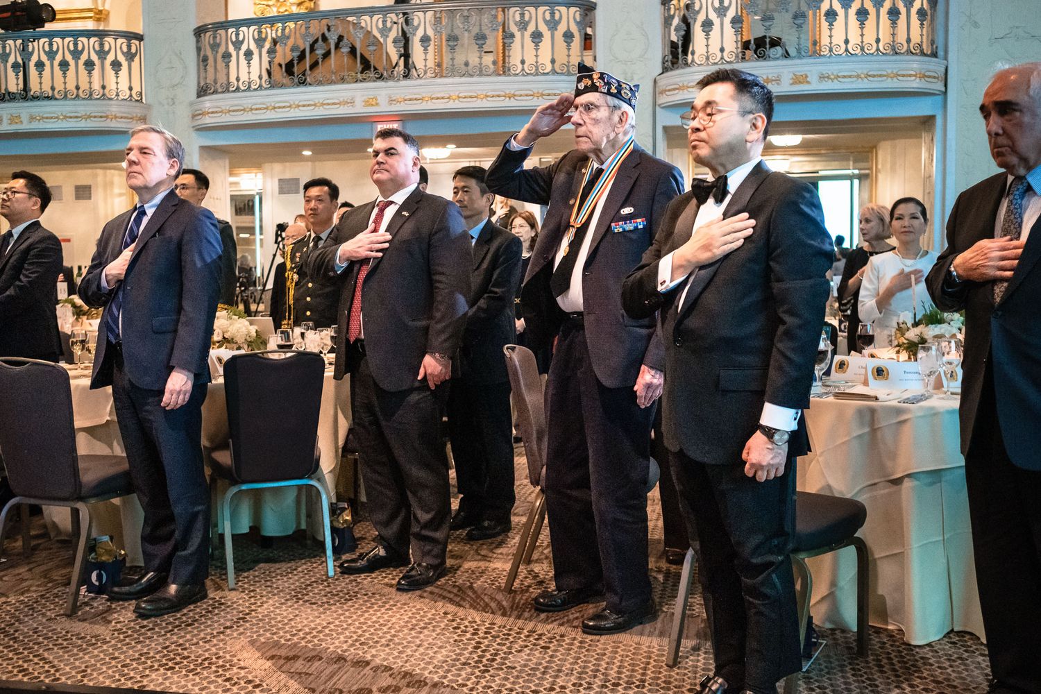 Men in formal attire stand at attention during a patriotic ceremony at an elegant banquet hall event.