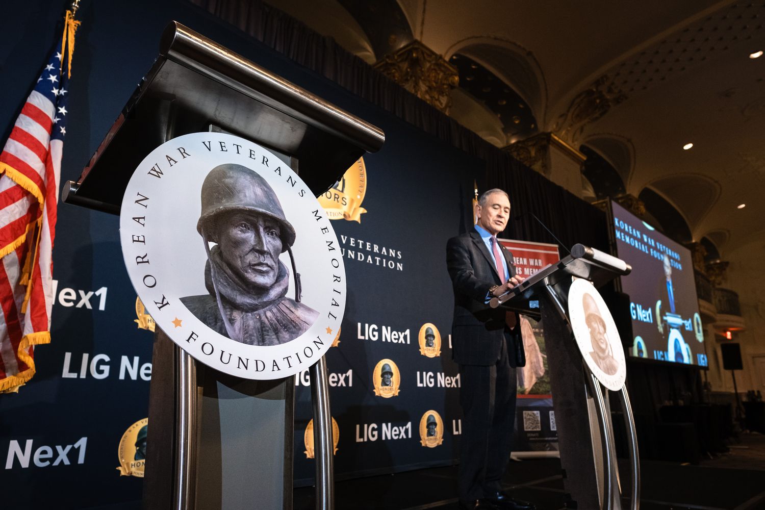 Speaker at podium during Korean War Veterans Foundation event with LIG Nex1 sponsorship banners.