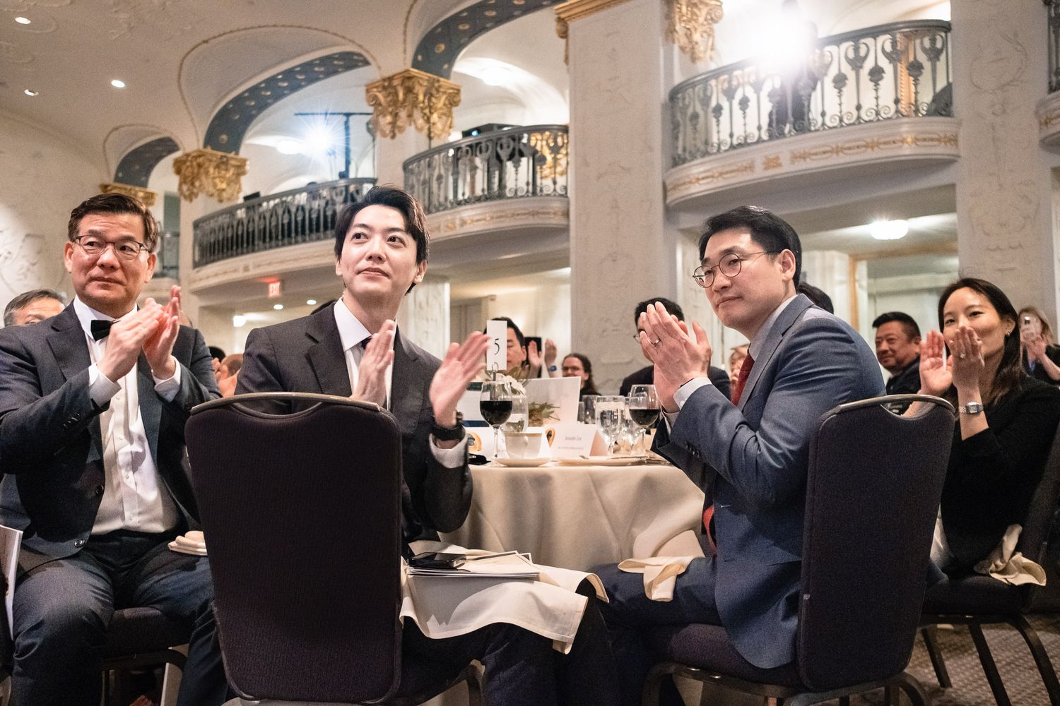 Business professionals in suits applauding at a formal banquet event in an elegant ballroom setting.