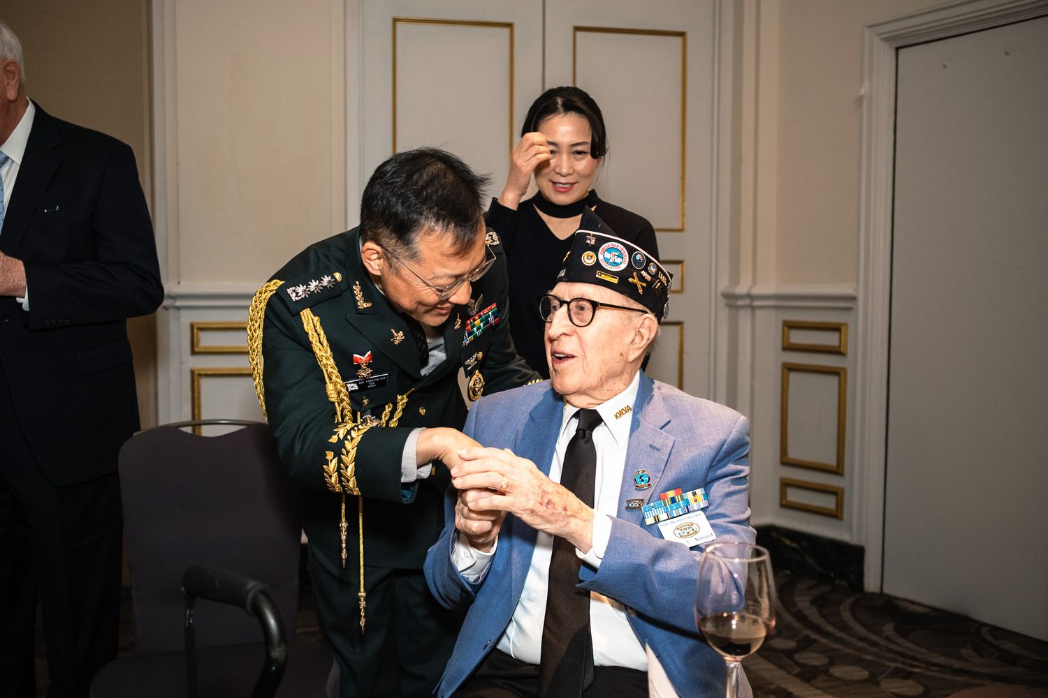 Military officer shakes hands with a seated veteran in a blue blazer at a formal event.