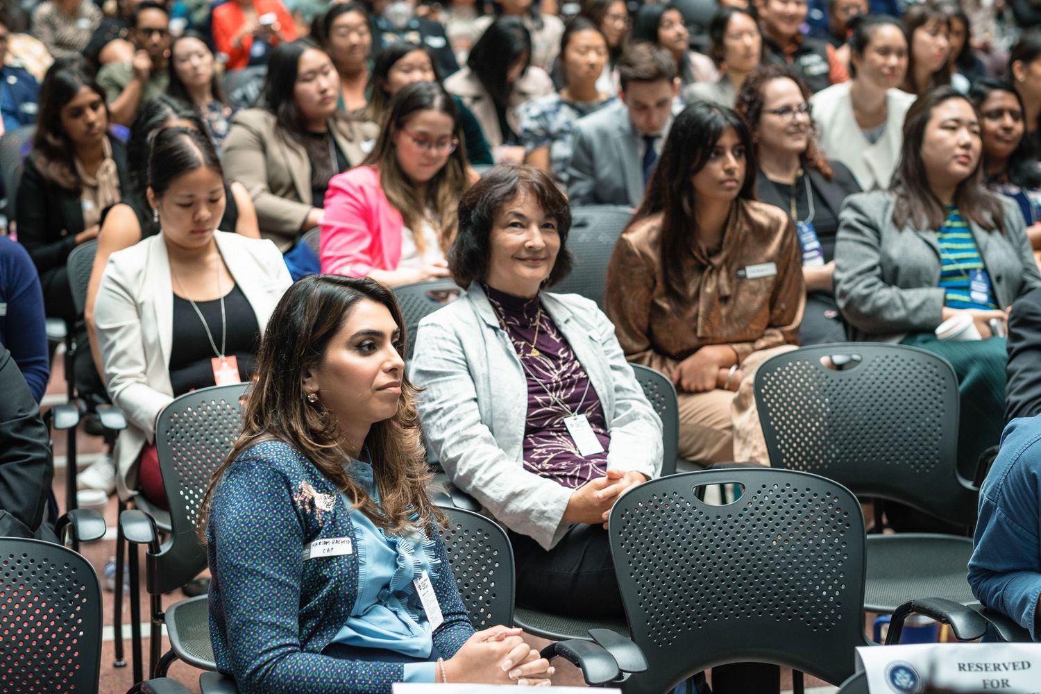 Diverse audience of women attending a professional conference, seated in rows of chairs in a large auditorium.