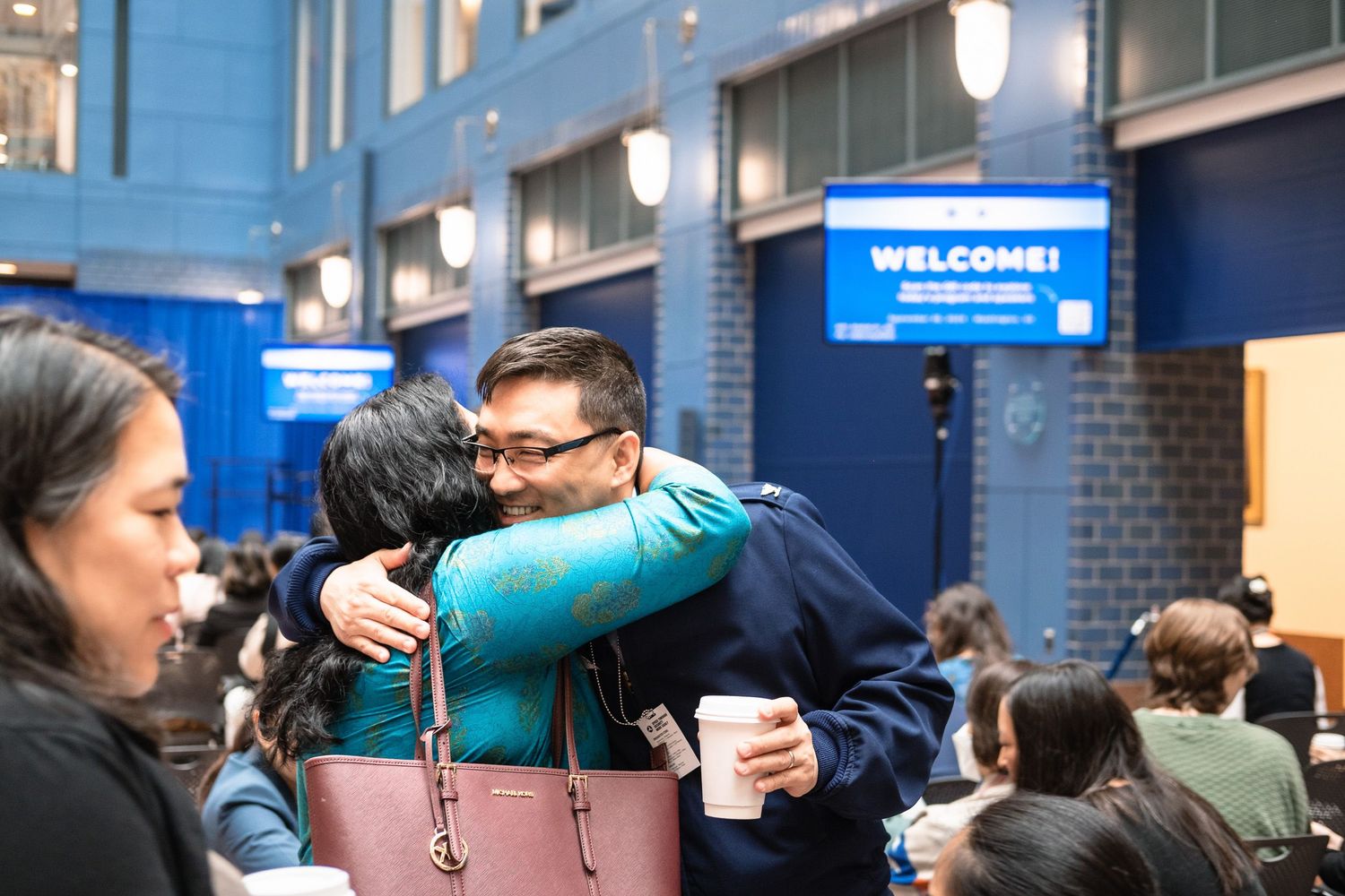 Two people share a warm hug at a welcome event inside a blue-decorated venue with attendees in the background.