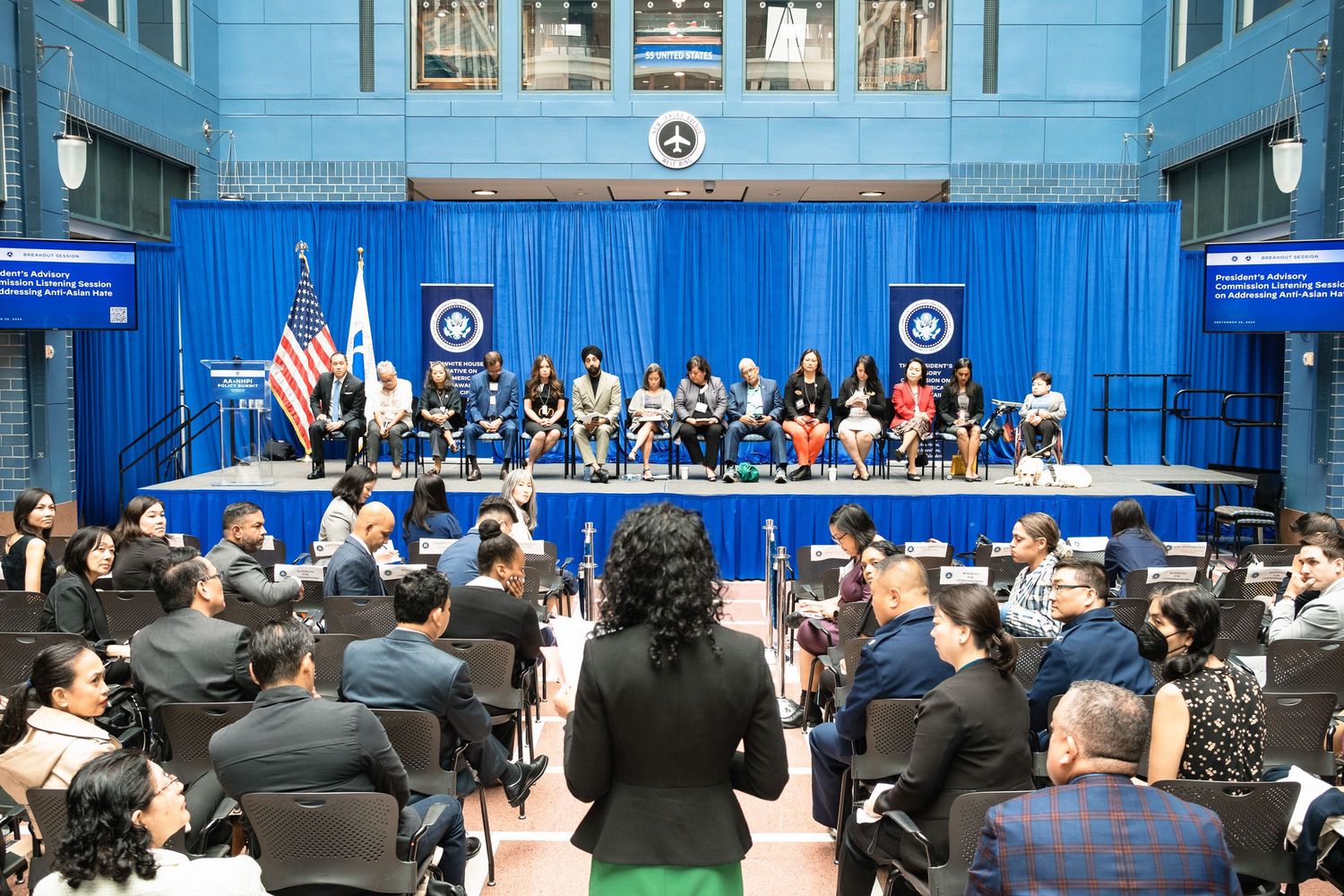 Formal government ceremony with officials seated on stage before an audience in a blue-draped hall with US flags.